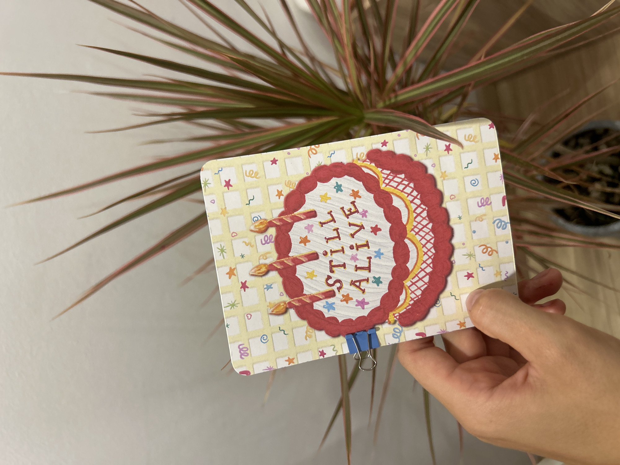 A person's hand holds a rectangular birthday card in front of a Dracaena plant. The card has a yellow checkered background with colorful confetti and stars. A drawing of a red-frosted cake with three lit candles is on the front of the card. The words "STILL ALIVE" are written on the cake in a festive font. A blue binder clip is attached to the left edge of the card. The background is a plain, light-colored wall and the plant's foliage extends into the frame. The image was taken in Chang Phueak, Thailand.