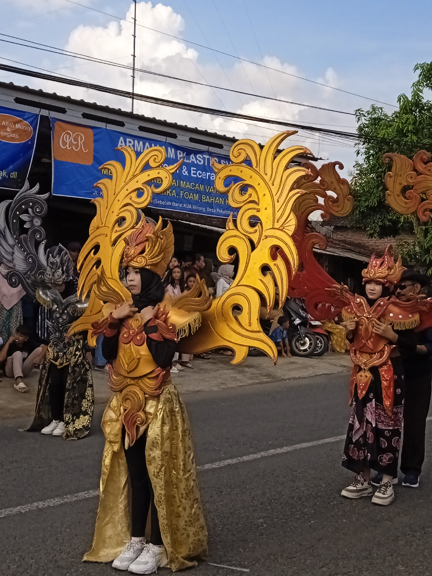 The image captures a street scene in Winong, Indonesia. Three individuals are costumed in ornate, colorfully embellished garments reminiscent of traditional Indonesian attire. The costumes feature prominent wing-like backpieces: one in black and silver, another in gold, and a third in red and gold. Each individual is wearing distinct headwear. The person in the gold costume is wearing a hijab. The other two are wearing matching shoes with thick soles.
In the background, a group of spectators is visible, along with a parked motorcycle. Overhead, electrical wires are strung. A blue banner displays text reading "ARMAM PLASTIC," suggesting a local business, and includes the phrase "(Sebelah Barat KUA Winong, Desa Pekalong.n)".