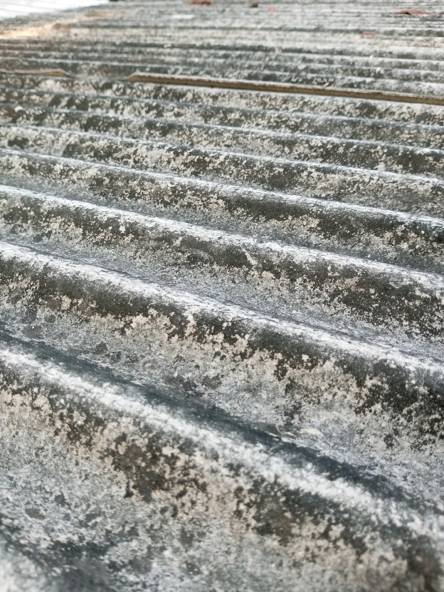 The image shows a close-up, angled view of corrugated roofing material. The material appears aged and weathered, with a mottled texture of dark gray and white, likely from moss, lichen, or dirt accumulation. The corrugations create a ribbed pattern that recedes into the distance, with a shallow depth of field causing the foreground to be in sharper focus than the background. The lighting suggests an outdoor environment, possibly under daylight. There are no people or specific identifiable landmarks visible. The description is limited to what can be observed in the image itself and does not include any external information or speculation about location.