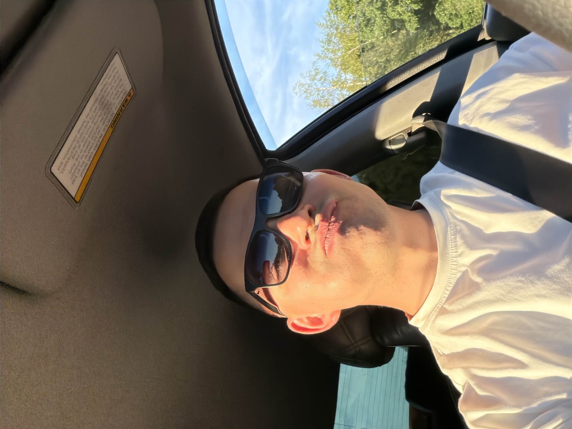 An individual is captured in a close-up selfie or portrait while seated inside a vehicle, identified as a car in Yus'ki, Russia. The person, appearing to be male, is seen from the chest up, with their head tilted to the right. They are wearing dark sunglasses and a white t-shirt, secured by a seatbelt across their chest. Their lips are pursed, suggesting a playful or affectionate expression.
The setting is bright and sunny, indicated by clear blue skies with scattered clouds visible through what appears to be a sunroof or open window. Green foliage from trees can be seen outside the car, suggesting a natural outdoor environment. Golden sunlight illuminates the person's face and shirt, hinting at either early morning or late afternoon light.
Visible within the car's interior is a sun visor with a prominent "WARNING" label. Despite some text being obscured or garbled by the image quality and angle, key phrases like "DEATH OR SERIOUS INJURY can occur" are discernible. The label also mentions "SAFEST place for children," "airbag," "seat in the front," and "SEAT BELTS and CHILD RESTRAINTS," advising to refer to a "manual for further information." The image is rotated, giving a unique perspective to the scene.