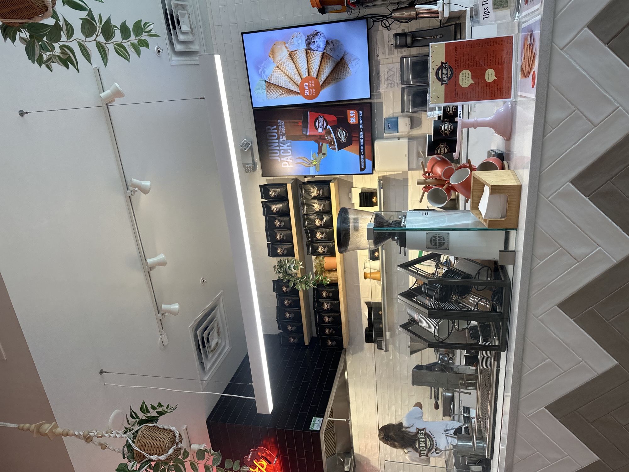 Here's a detailed description of the image:
**Setting:** The image appears to be taken inside a Lukumades dessert shop. The setting has a modern aesthetic with white walls, herringbone tile details, black tiled accents, and track lighting. A neon sign is partially visible near the lower edge of the image.
**Objects and Elements:**
* **Signage:** Digital displays advertise Lukumades gelato and the "Junior Pack" featuring donuts and drinks. Printed menus and signage are visible near the counter.
* **Products:** Packaged goods are stocked on shelves.
* **Equipment:** A coffee grinder, espresso machine, and other food preparation equipment are situated on the counter.
* **Accessories:** Mugs, napkins, and serving utensils are arranged on the counter space.
* **Decor:** Plants in hanging baskets adorn the interior.
**People:**
* An employee, wearing a "Lukumades" branded shirt, is visible working behind the counter.
**Action and Interactions:**
* The employee is likely engaged in preparing or serving food or drinks.
**Inferences:** The setting suggests a food service business catering to a modern and possibly family-oriented clientele. The presence of various equipment and products indicates a functioning retail environment.
**Location:** Based on the store name, it is likely that this photo was taken in Jacksonville Beach, FL, United States of America.