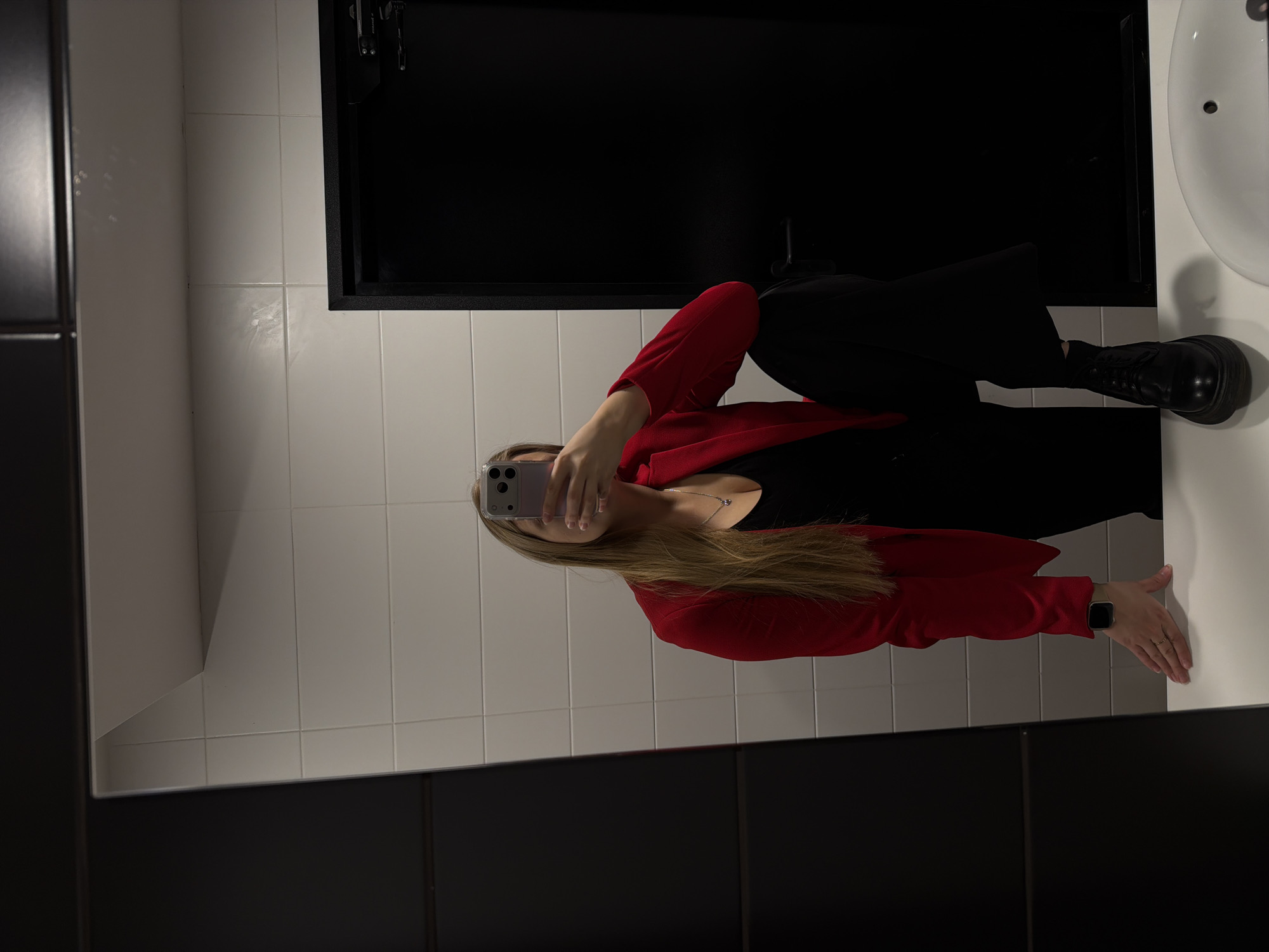 A female is reflected in a mirror, holding a smartphone to obscure her face while taking a selfie. She is wearing a black top, black pants, a red blazer, and black boots with thick soles. She is standing in a bathroom with white tiled walls and black fixtures, including a door frame and cabinetry. A white sink is partially visible on the right. There is no indication of the location in Izhevsk, Russia.