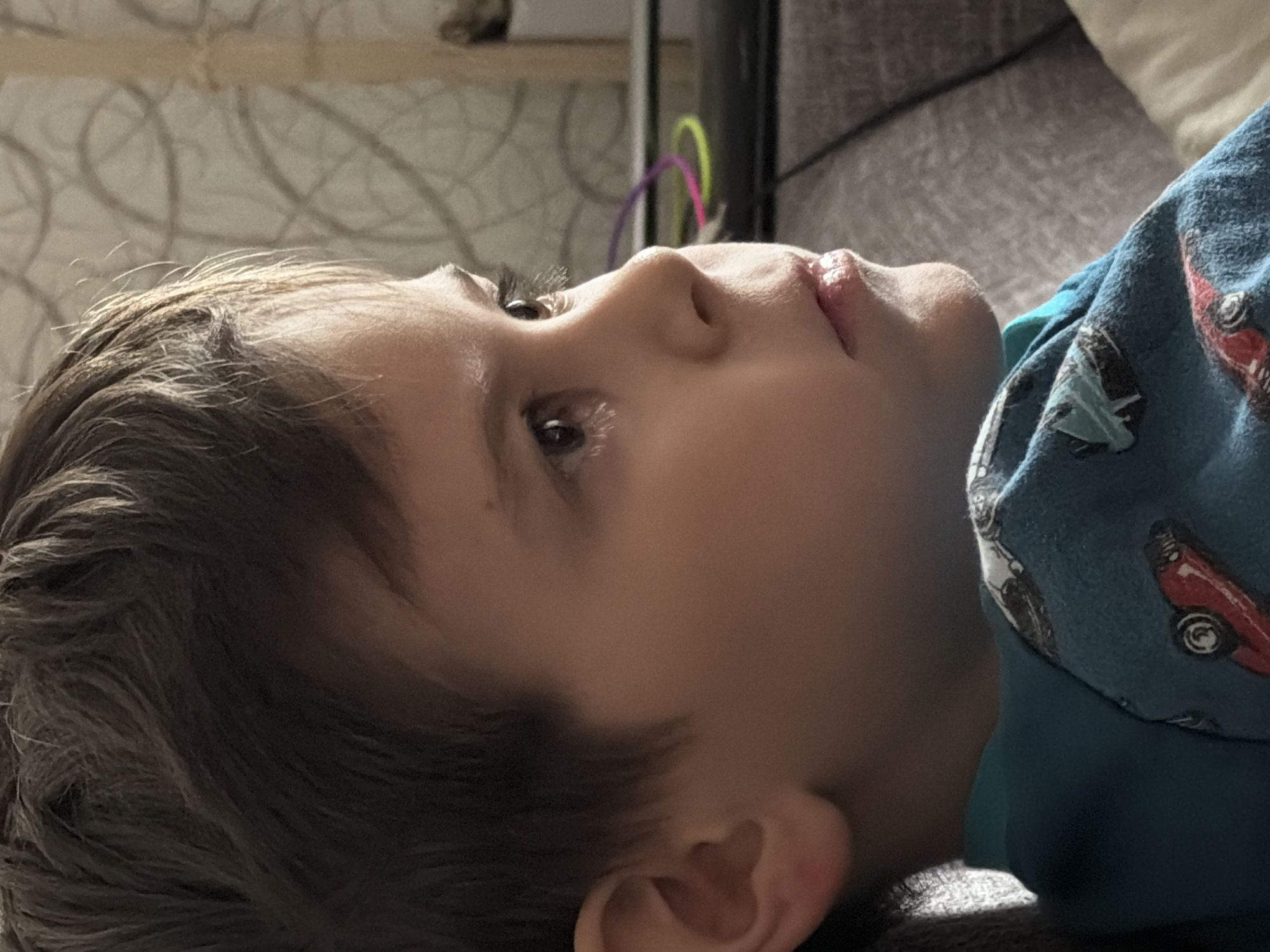 The image presents a close-up, profile view of a young child, likely a boy, lying down indoors in Perm, Russia. The child, with brown hair, is looking upwards and to the right of the frame with a contemplative or observant expression, his lips slightly parted. He is wearing a blue long-sleeved top patterned with various vintage cars, including red and white vehicles.
The background features a light-colored wall with a subtle, swirling textured pattern. Behind the child's head, parts of soft furnishings, possibly a grey sofa or bed, are visible. In the upper background, faint outlines of colorful, curved objects, resembling parts of a child's toy or activity arc, can be seen. The lighting is soft and ambient, characteristic of an indoor setting during the daytime. No specific activity beyond lying down and observing is depicted. There is no visible text in the image.