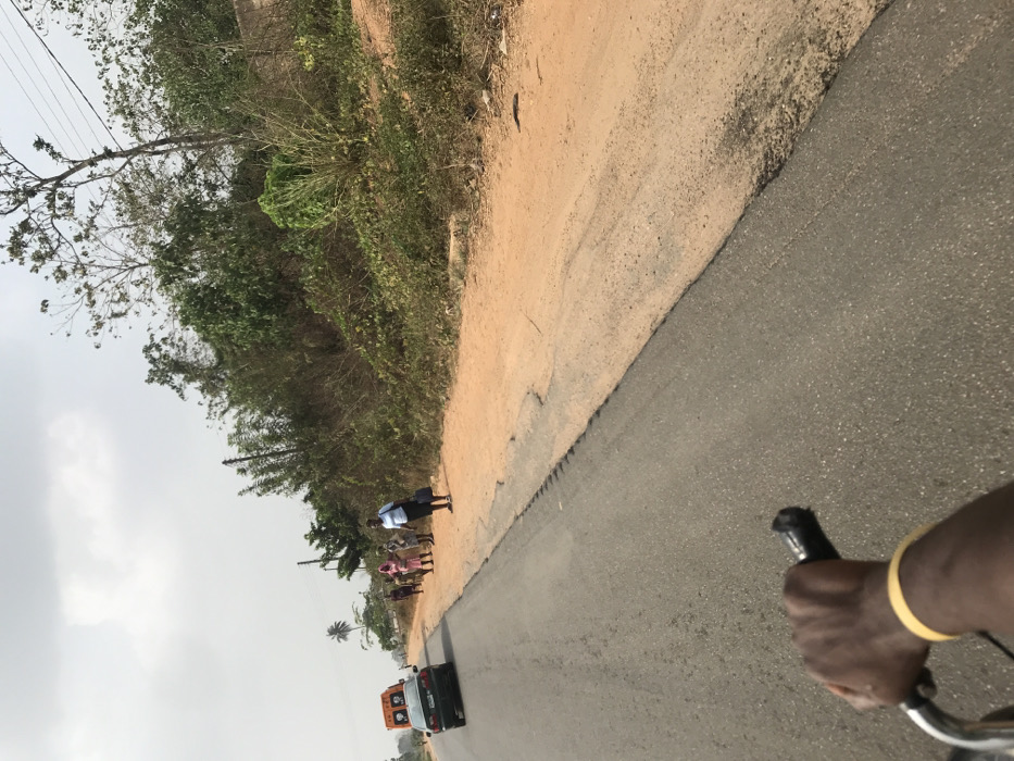 The image depicts a roadside scene in Ogbese, Nigeria, from the perspective of an individual riding a bicycle or motorcycle, evidenced by a dark-skinned right hand gripping a black handlebar in the lower foreground, wearing a yellow wristband. An asphalt road, exhibiting cracks and uneven edges, occupies the left and central portions of the frame, alongside a wide, reddish-brown dirt shoulder to its right.
In the mid-ground, five individuals are visible on the dirt shoulder. The foremost person is a woman in a light blue top and dark skirt, carrying a dark bag, oriented to her left. Behind her, four additional individuals, primarily women in casual clothing, stand further down the road.
A dark sedan travels away from the viewer on the asphalt road in the mid-ground. Behind it, a light-colored vehicle with an orange or yellow roof attachment and red/white markings on its rear is partially visible, also moving away.
The environment features dense green and brown vegetation, including bushes, trees, and some bare-branched trees, lining the right side of the road. Power lines are visible running through the foliage. A single palm tree stands in the distant left. The sky overhead is overcast and light gray.
