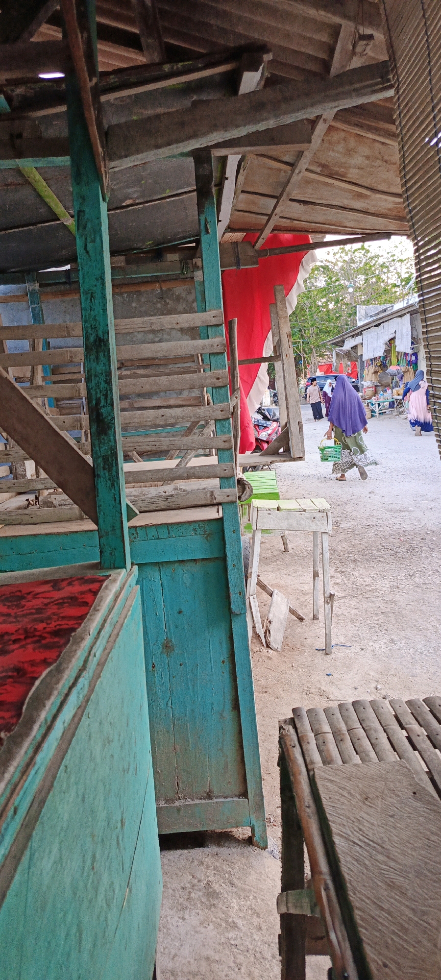 In a dusty, outdoor market setting, likely in Gayam, Indonesia, a person wearing a purple hijab and a patterned dress walks away from the viewer, carrying a green plastic basket. They are positioned in the middle ground of the image, towards the right side. Behind them, a red and white cloth hangs from a wooden structure, partially obscuring a glimpse of trees and sky. Further in the background, other market stalls with various goods and people can be seen. The foreground features the weathered, turquoise-painted wooden structure of a market stall, with wooden planks and a red-covered surface visible. Another bamboo bench is also present in the foreground, to the right. The ground is covered in dirt and gravel.