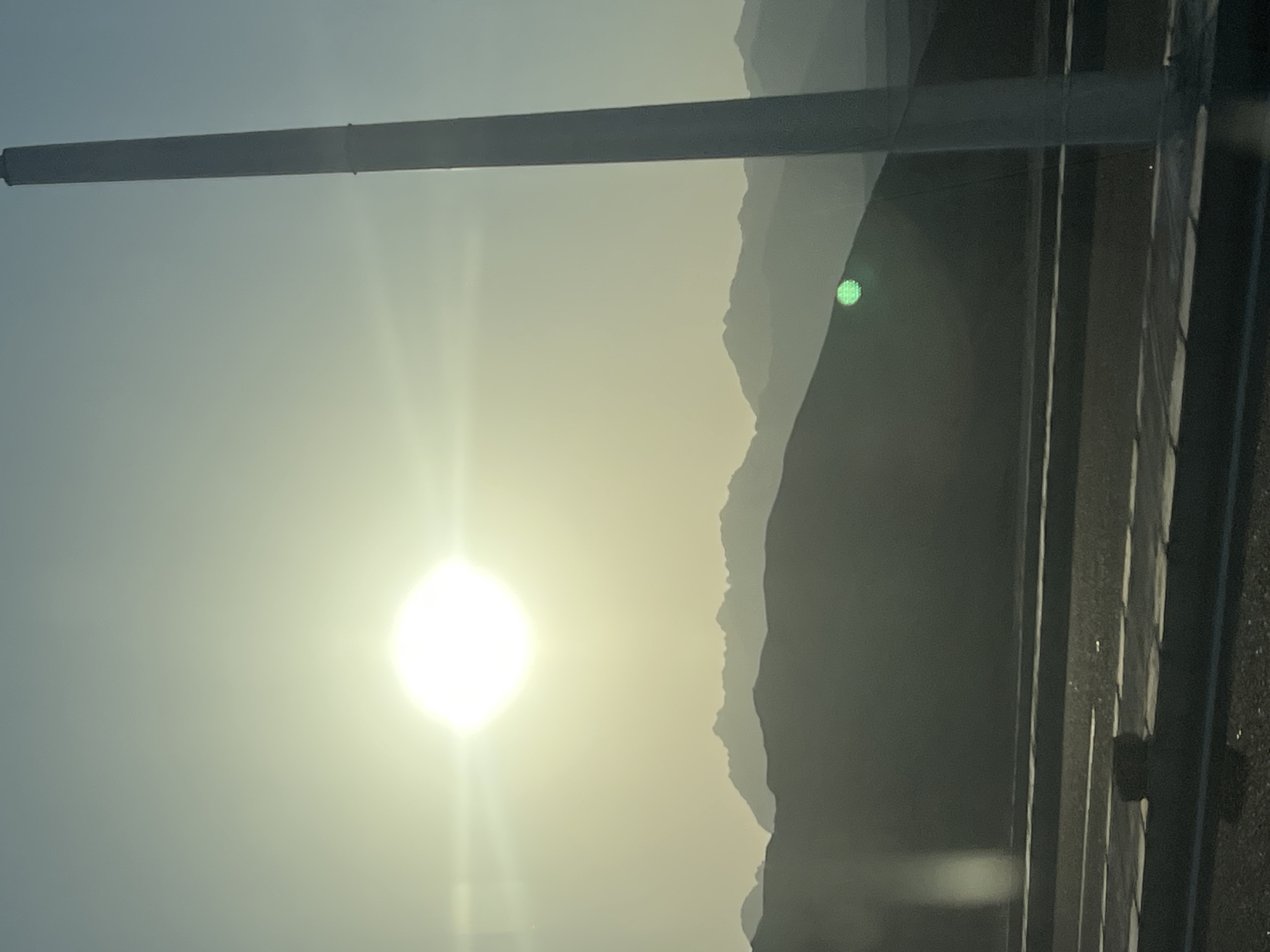 A bright, hazy sun dominates the upper left quadrant of the image, casting a soft glow over a silhouetted mountainous landscape in the background. A tall, slender, white pole stands vertically on the right side of the frame, extending from the bottom edge towards the top. In the mid-ground, a road with white markings is visible, and a dark, shadowy object is present on the road surface in the lower left. The overall impression is a vast, outdoor scene, likely taken during daylight, with a focus on natural elements and a man-made structure. The image evokes a sense of an open environment, potentially a highway or roadside, overlooking distant mountains under a bright sky. The presence of the sun and the hazy atmosphere suggest it could be early morning or late afternoon.