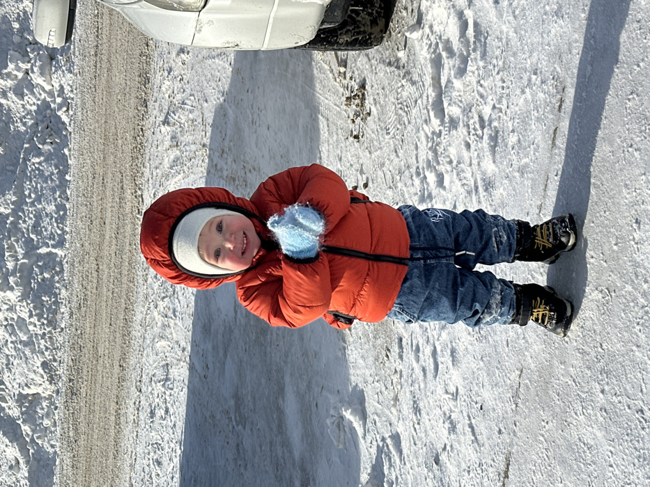 A young child, appearing to be a toddler, stands centrally on a snow and ice-covered surface in Perm, Russia. The child wears a bright orange-red hooded puffer jacket over a white beanie, blue denim jeans, and black winter boots with yellow laces. Both hands are covered in light blue, fluffy mittens and held together at the chest. The child's face is visible, displaying a smiling expression. The ground consists of packed snow, patches of ice, and scattered dark debris. To the right, the lower front quarter and wheel of a dirty, light-colored vehicle are partially visible. In the background, a large snowdrift is present, indicating a cleared area, behind which a darker, slush-covered surface is discernible. The scene is brightly illuminated, casting clear shadows on the snow.