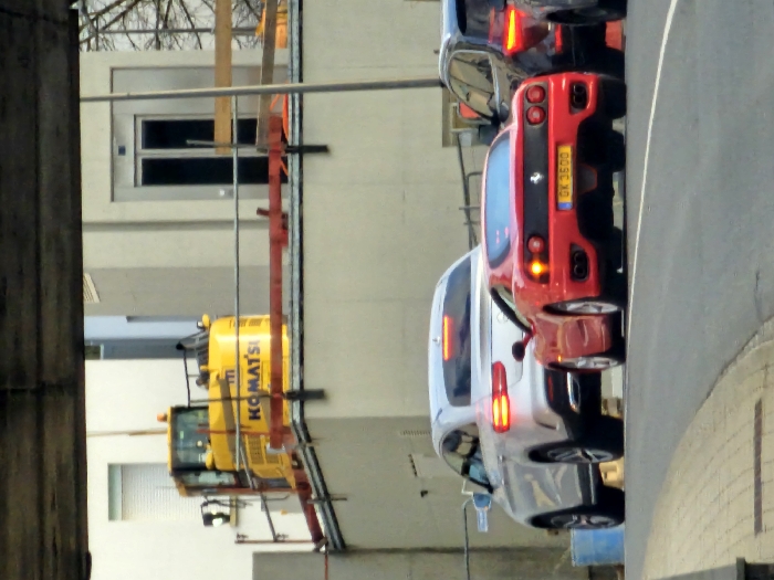 An urban street scene in Schifflange, Luxembourg, is depicted from under an overhead concrete or metal structure. Three vehicles are positioned on a paved road, facing away from the viewer. The central vehicle is a red Ferrari 360 Modena, displaying Luxembourg license plate "GX 3600" and illuminated taillights. To its left and slightly behind is a silver Mercedes-Benz GLE Coupe, also with illuminated taillights. A dark-colored SUV is partially visible on the far right, behind the Ferrari, showing its left rear and illuminated taillights. A grey paved sidewalk borders the left side of the road, and a blue barrel is partially visible near the sidewalk on the left. In the background, on an elevated platform or building roof, a large yellow Komatsu excavator is visible, surrounded by red and silver construction barriers. Behind the excavator, several light-colored buildings are present, one featuring a prominent external elevator shaft with dark doors and another with a white window blind. No individuals are visible in the scene.