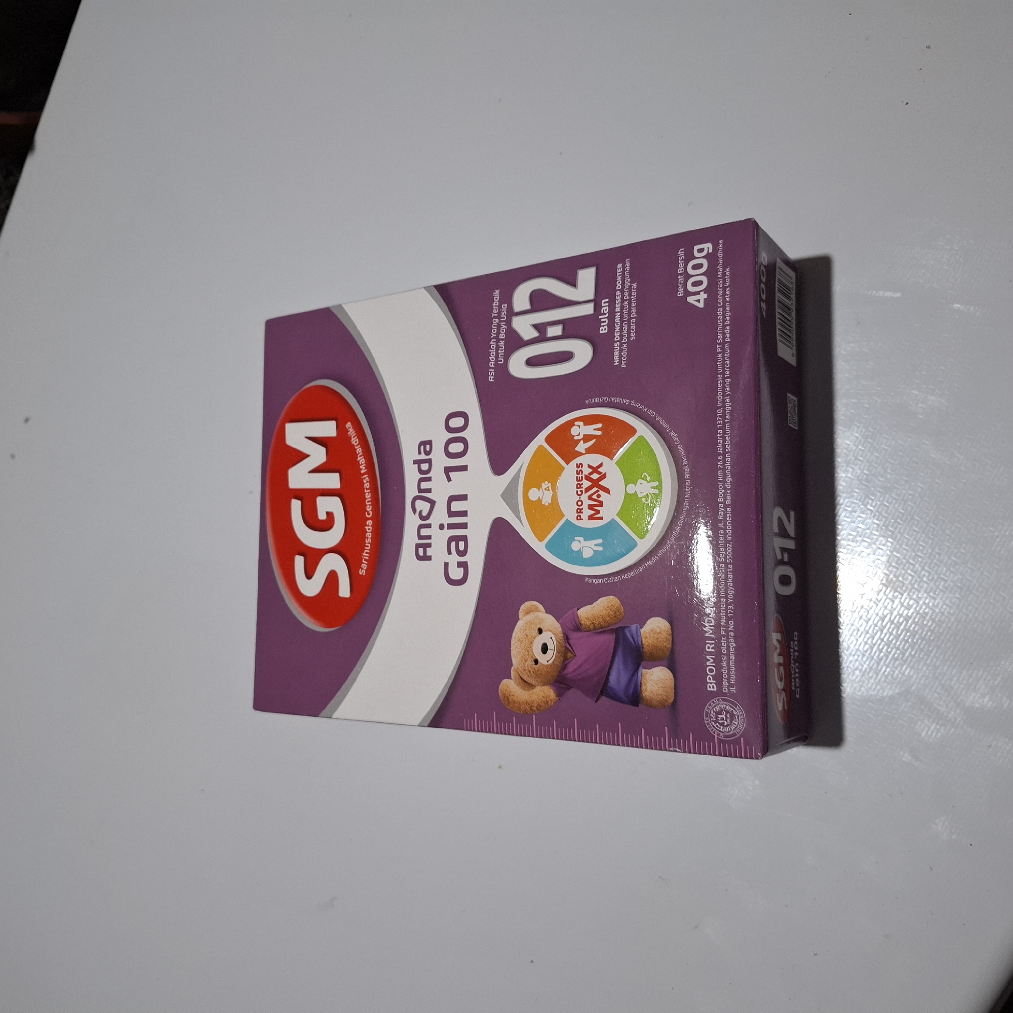 The image shows a close-up, slightly angled view of a SGM Ananda Gain 100 formula milk box, predominantly purple and white, resting on a plain, light-colored surface, possibly a table. The box features branding, product information, and graphics, including a teddy bear illustration and a circular graphic with icons. The lighting is soft and diffused, casting minimal shadows. The background is out of focus, showing a blurred dark area at the top right, suggesting an indoor setting. There are no people visible in the image, and no specific location in Reudeuep, Indonesia can be determined from the visual information provided.