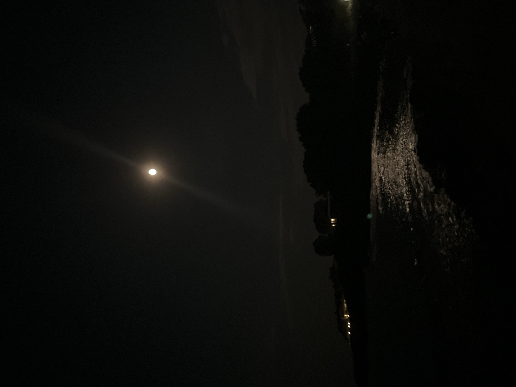 The image shows a nocturnal scene with a full moon illuminating a body of water. In the foreground, dark rocks line the shore. The water's surface reflects the moonlight, creating a shimmering effect. In the mid-ground, a dark silhouette of trees and vegetation is visible along the coastline. Behind the trees, a few scattered lights suggest the presence of buildings, possibly houses or resorts. The overall ambiance is serene and tranquil, characteristic of a peaceful night by the sea. The location appears to be the coast of Lozenets, Bulgaria, a Black Sea resort town known for its beaches and natural beauty. The image captures the charm of Lozenets at night, with the moonlit sea and the subtle glow of distant lights painting a picturesque scene.