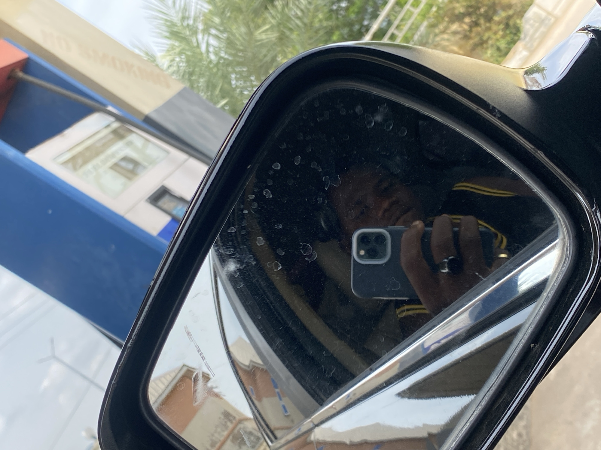 The image is a close-up, angled shot of a car's side mirror, reflecting a person holding a smartphone. The mirror itself is dirty with water spots. In the reflection, a dark-skinned individual is visible, wearing a black shirt with yellow stripes and a ring on their left index finger. They are holding a dark-colored smartphone, likely an iPhone, to take the picture.
The background reflected in the mirror shows elements of an urban environment, possibly a gas station or a commercial building, with blue and white signage. The presence of palm trees suggests a tropical or subtropical climate. The bright daylight indicates it is daytime. The scene is likely located in Jalingo, Nigeria, given the context provided. The overall impression is a casual self-portrait taken in an outdoor setting within a Nigerian city.