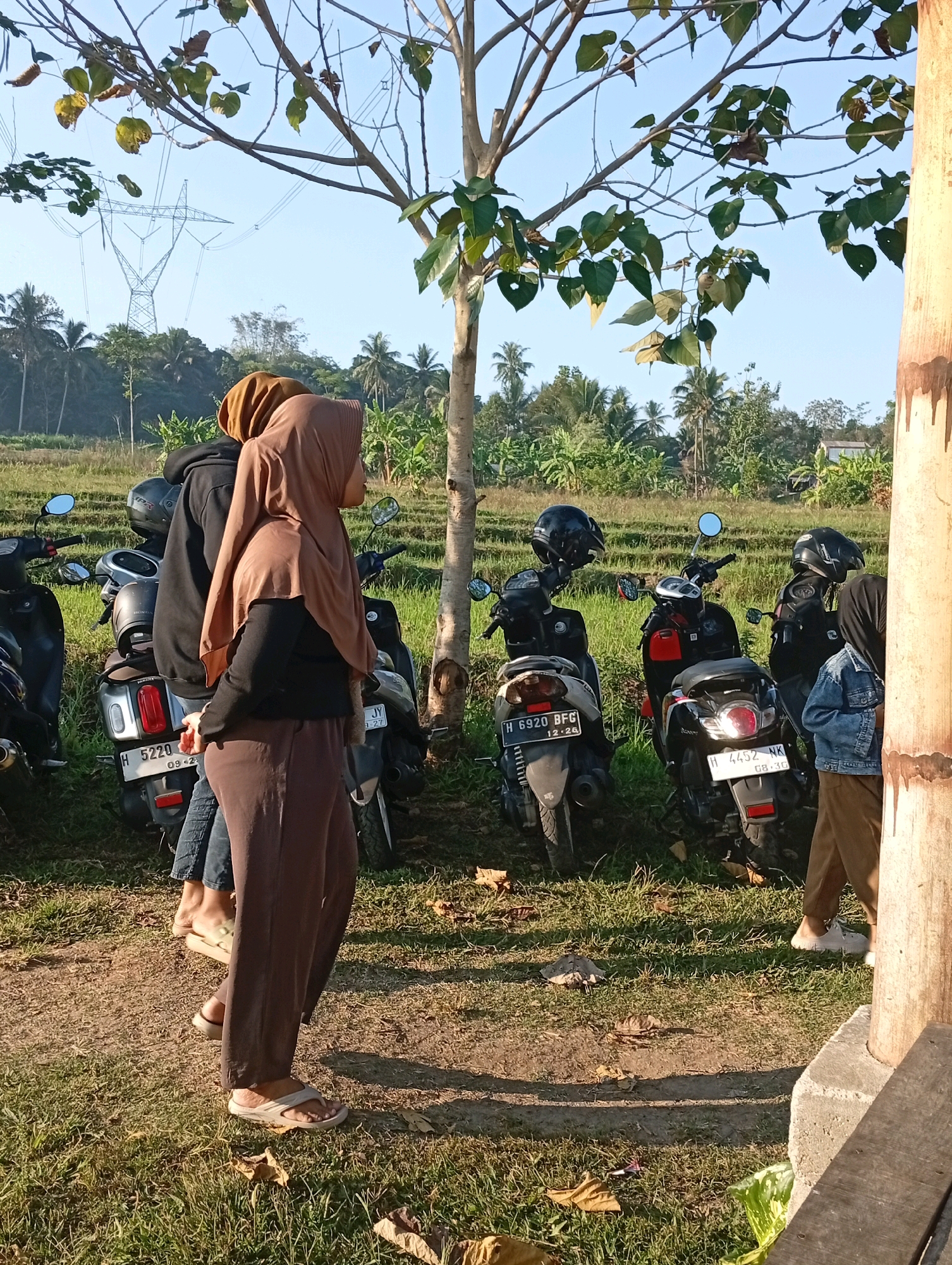 A group of young individuals, likely friends, are gathered around parked motorcycles in a rural outdoor setting. The scene appears to be a grassy field with a few trees and agricultural land in the background, suggestive of a location outside of a dense urban area. The presence of tall power transmission towers in the distance hints at proximity to a utility corridor, possibly on the outskirts of a city or town. Given the tropical vegetation and architectural style of the buildings in the background, this scene could be located in the vicinity of Salatiga, Indonesia, a city known for its agricultural landscapes and mountainous terrain. The individuals are dressed casually, with some wearing headscarves, indicating cultural context. Their interaction suggests a moment of pause or preparation before or after a journey, perhaps a leisure ride or a visit to a rural spot. The overall atmosphere is relaxed and informal, capturing a slice of everyday life in this Indonesian locale.