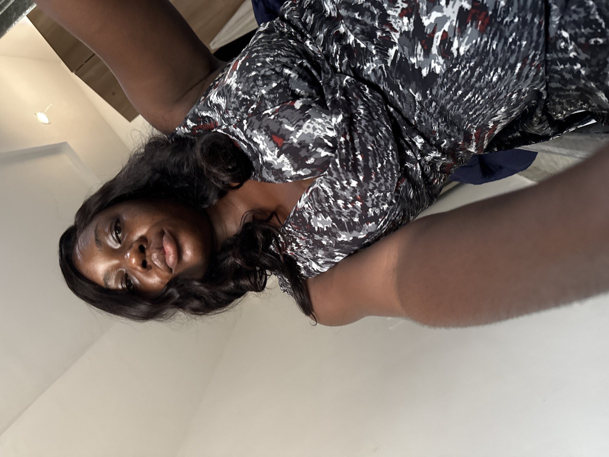 The image displays a close-up, upward-angled shot of an individual, likely a woman, in an indoor setting, possibly in Iddo Tudunwada, Nigeria. The subject has dark skin and long, wavy, dark hair that frames her face. She looks directly into the camera with a calm expression, her lips slightly upturned in what appears to be a subtle smile.
She is dressed in a patterned top or dress featuring an abstract design composed of black, various shades of grey, white, and occasional flecks of red. Her bare arms extend towards the camera, suggesting she is holding the device or positioning herself for the shot.
The background is predominantly a plain, light-colored wall or ceiling, indicating an interior space. A small, round light fixture with a visible bulb is present in the upper left area of the frame, providing artificial illumination. A dark blue fabric is also partially visible behind her, possibly part of clothing or bedding. The perspective suggests the subject is lying down or reclining. There is no visible text in the image. The time of day cannot be determined from the indoor lighting.
