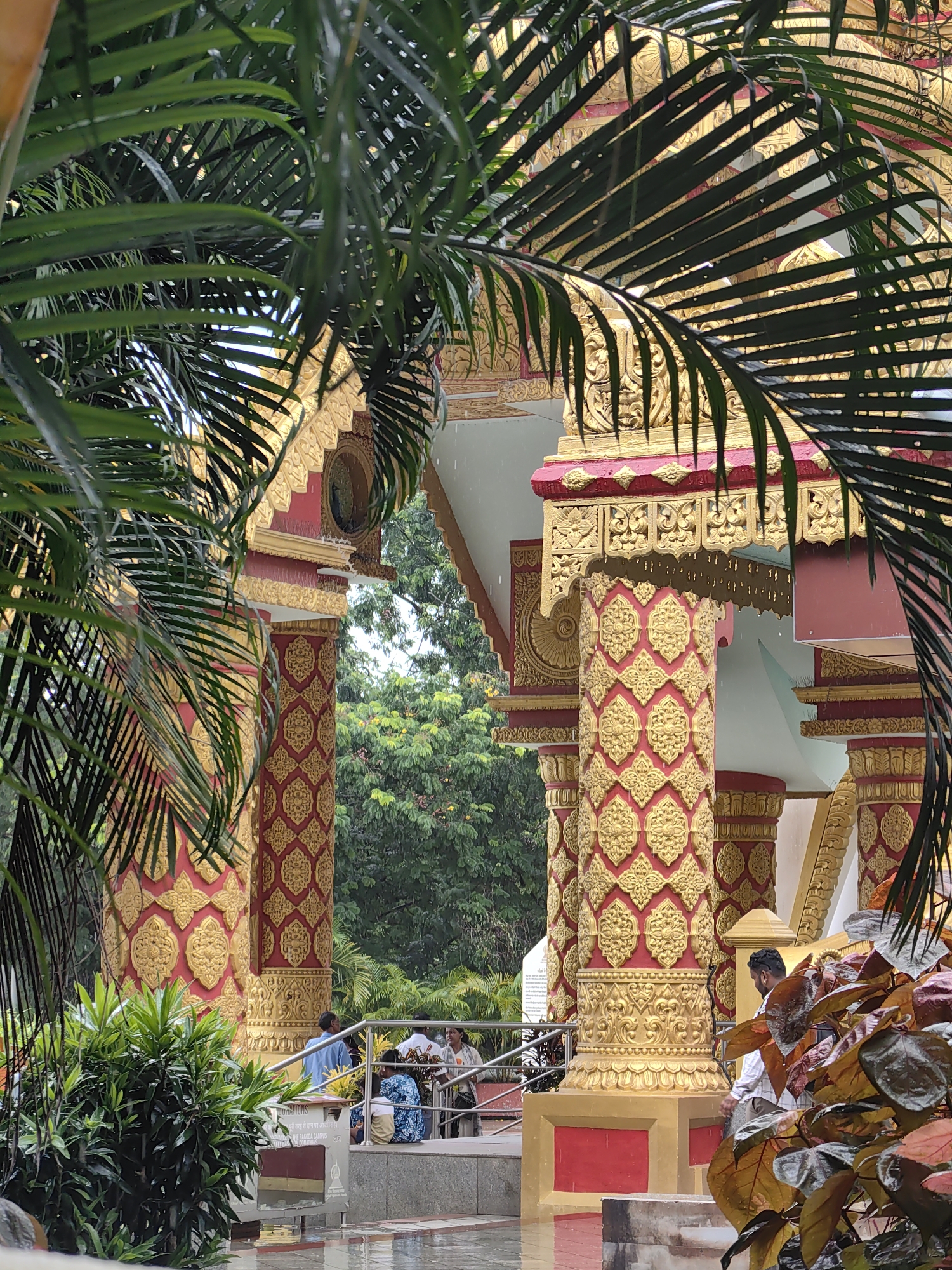 The image depicts the exterior of a Buddhist temple, likely in Mumbai, India, characterized by its ornate red and gold pillars adorned with intricate floral patterns. Lush green palm fronds are prominently featured in the foreground, partially obscuring the view of the temple architecture. In the background, a dense canopy of trees suggests a park-like or natural setting. Several people are visible in the mid-ground, some standing near a railing and others seated on steps, appearing to be visitors or devotees. The ground is wet, indicating recent rain. The overall ambiance suggests a serene and spiritual atmosphere.