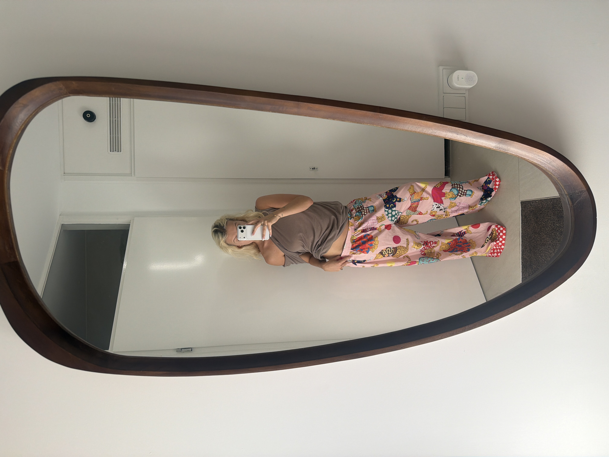 A blonde woman is taking a mirror selfie in a room with white walls. She is wearing an off-the-shoulder brown top and pink pajama pants with a colorful leopard print. Her left hand is on her hip, and her right hand is holding a smartphone. The room appears to be a hallway or entryway, with a closed door in the background and a light switch and a small white device on the wall to the right. The mirror itself is large and has a dark wooden frame with an irregular shape. The overall setting is modern and minimalist.