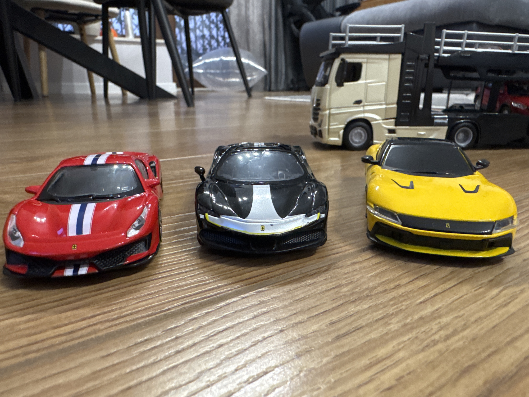 The scene depicts a collection of toy vehicles arranged on a wooden floor. Three Ferrari model cars dominate the foreground: one is red with white and blue stripes, the second is black with a silver stripe, and the third is yellow. Behind these, a larger toy truck with a car carrier trailer is partially visible. The background shows elements of a domestic setting, including furniture legs and a partially visible sofa. The location within the city of Plovdiv, Bulgaria cannot be determined based on the visual data provided.