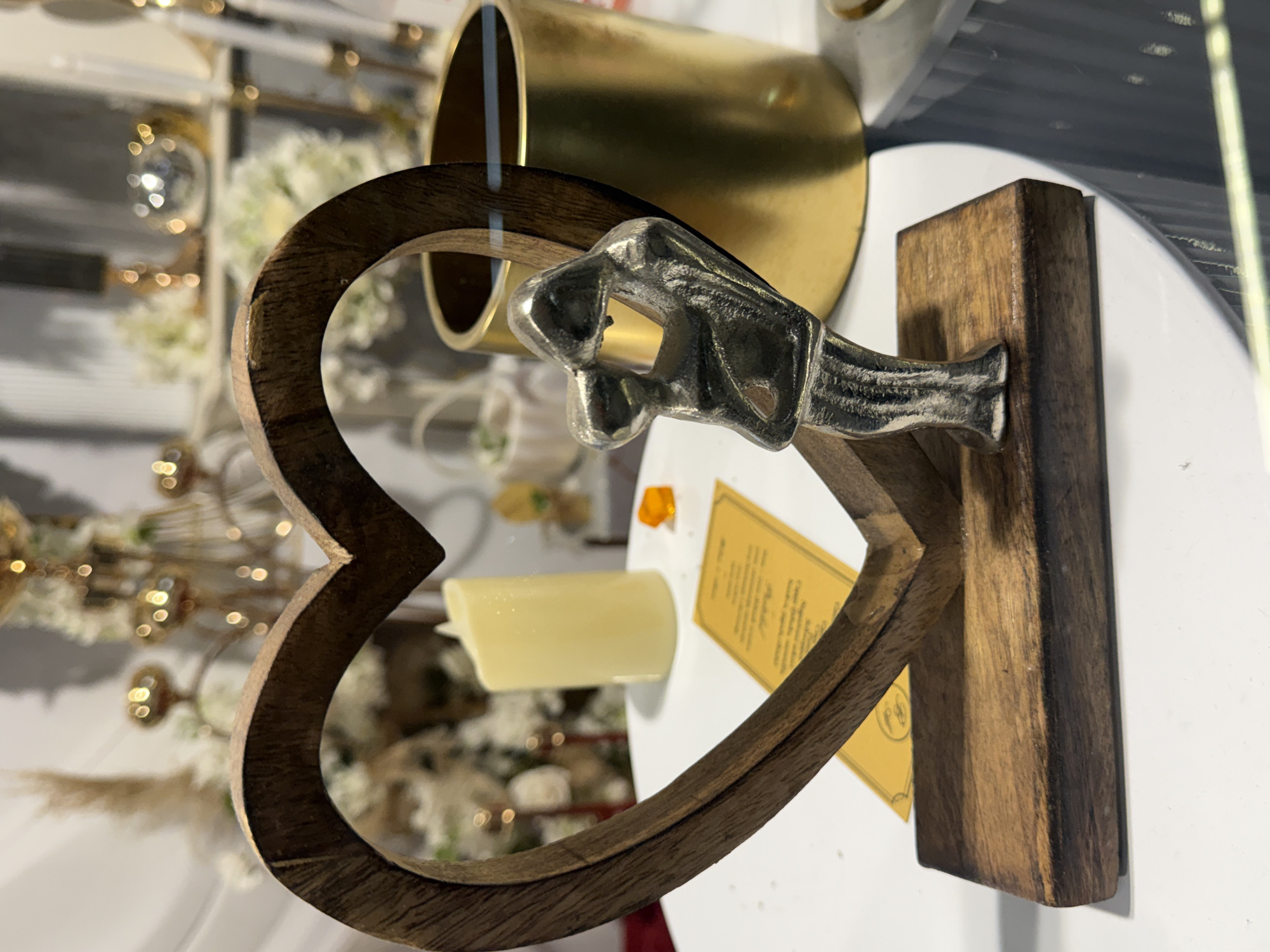 The image shows a close-up, eye-level, indoor shot of a decorative item. The main subject is a heart-shaped wooden frame with a small, silver figurine of a person with their arms raised in front of them, positioned on a wooden base. The figurine is inside the heart frame. Behind the figurine, to the right, is a cylindrical gold-colored container. To the left of the figurine and inside the heart, is a pale yellow, cylindrical candle. A yellow tag with text is placed on the surface in front of the wooden base. The background is blurred, showing hints of decorative elements like flowers and metallic objects, suggesting a retail or display setting. The scene does not provide any information that would allow for determining its location in Ilidža, Bosnia and Herzegovina.