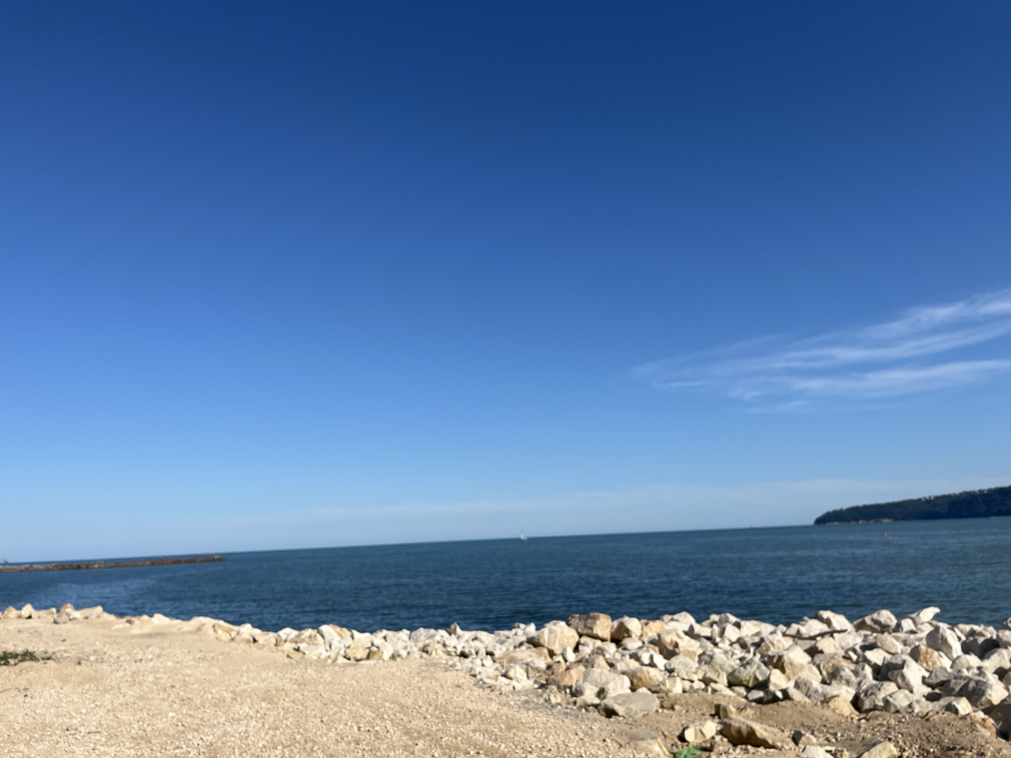 The image depicts a seascape with a clear blue sky. In the foreground, there is a rocky shoreline with sandy patches. The sea extends to the horizon, where a distant landmass is visible on the right. On the left side of the image, a man-made structure, possibly a pier or breakwater, juts out into the water. In the middle distance, a small sailboat can be seen. The overall scene suggests a coastal area with calm water and fair weather. This landscape is characteristic of the Varna Bay area in Varna, Bulgaria, known for its beaches and waterfront.
