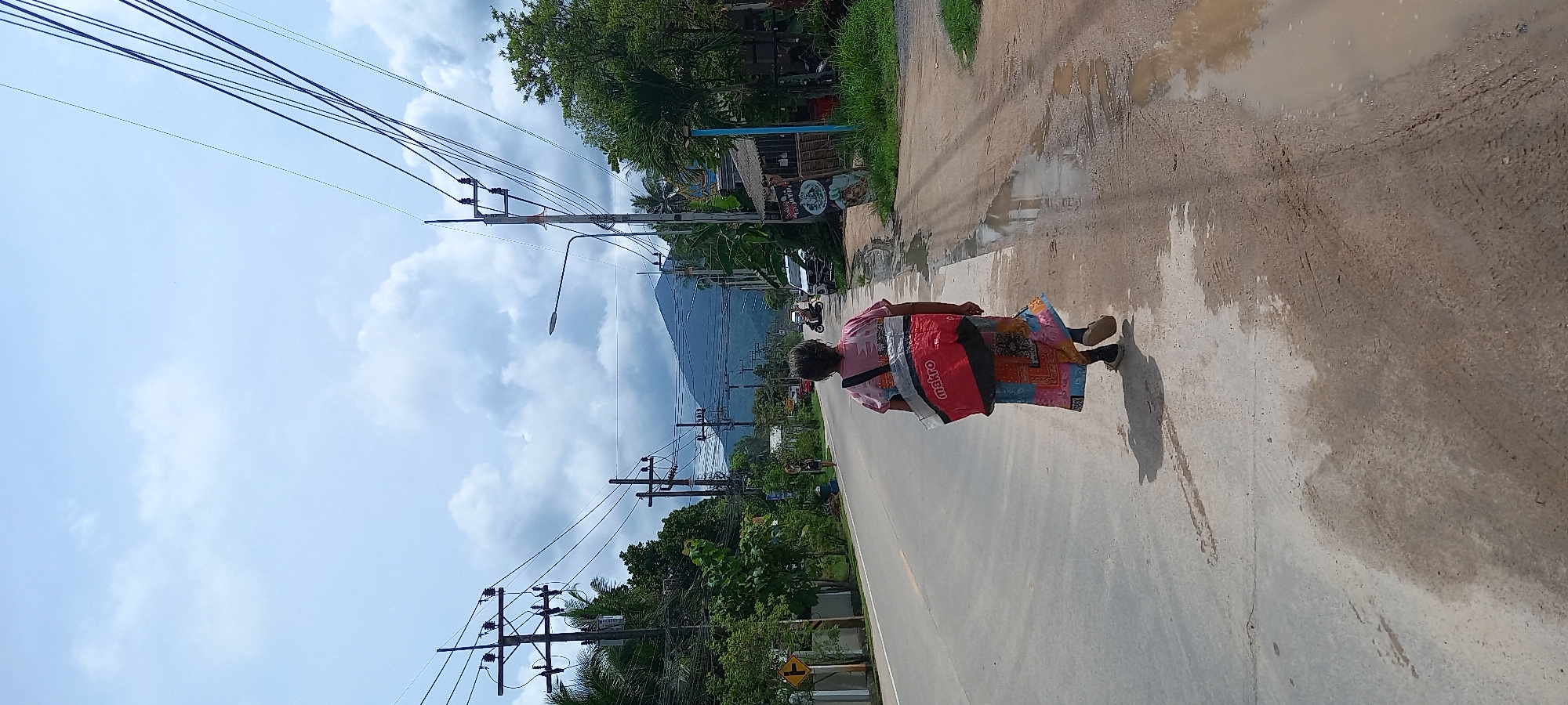 An elderly person wearing a red plastic poncho walks on the right side of a two-lane paved road in Thong Sala, Thailand. The road has patches of standing water along the curb. Power lines and utility poles run along the left side of the road. Lush green trees and vegetation flank both sides of the road. In the distance, a mountain is visible. A few buildings are interspersed among the trees. The sky is blue with scattered clouds.