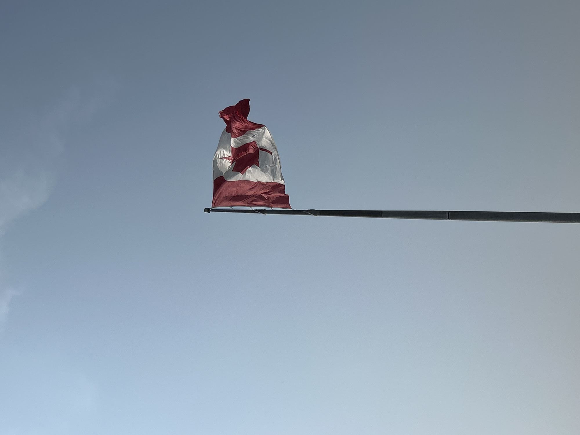 A Canadian flag waves on a flagpole against a pale blue sky with faint clouds. The flag is predominantly red and white, featuring the iconic maple leaf emblem. The flagpole is tall and grey, positioned on the right side of the image, with a portion of the flag extending to the left. The sky occupies the majority of the frame, creating a sense of open space. The image appears to be taken outdoors, under natural light.