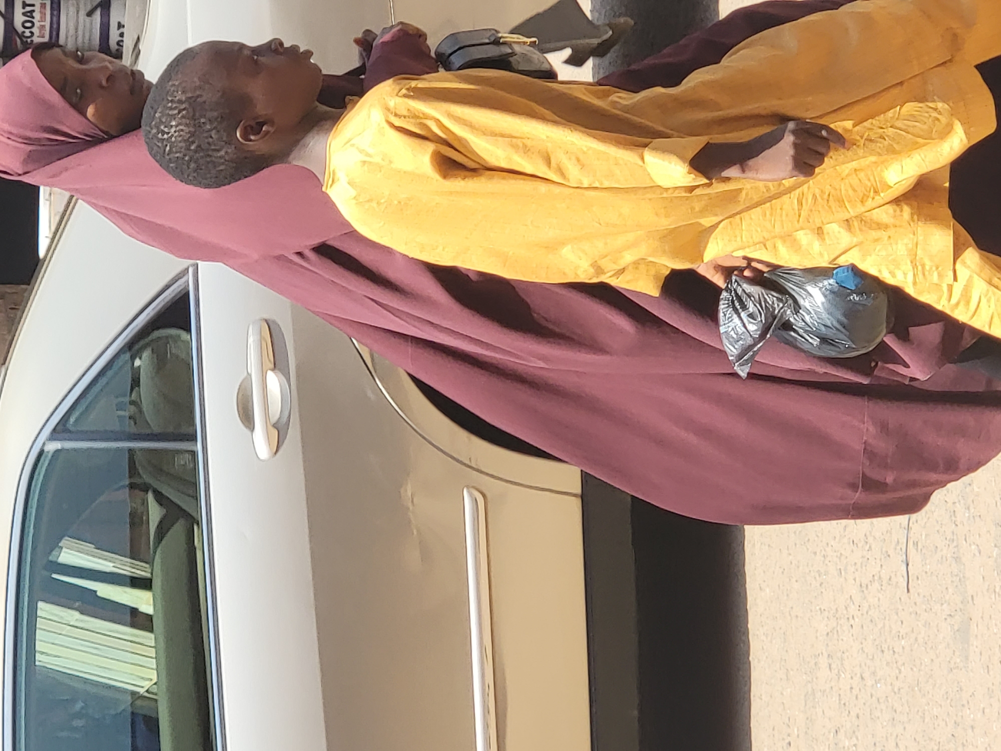 A young child in a yellow shirt, head resting on an adult wearing a mauve headscarf, sits next to a car on a street in Maiburiji, Nigeria. A shiny black plastic bag rests near the child's hand. The car is a light metallic color.
