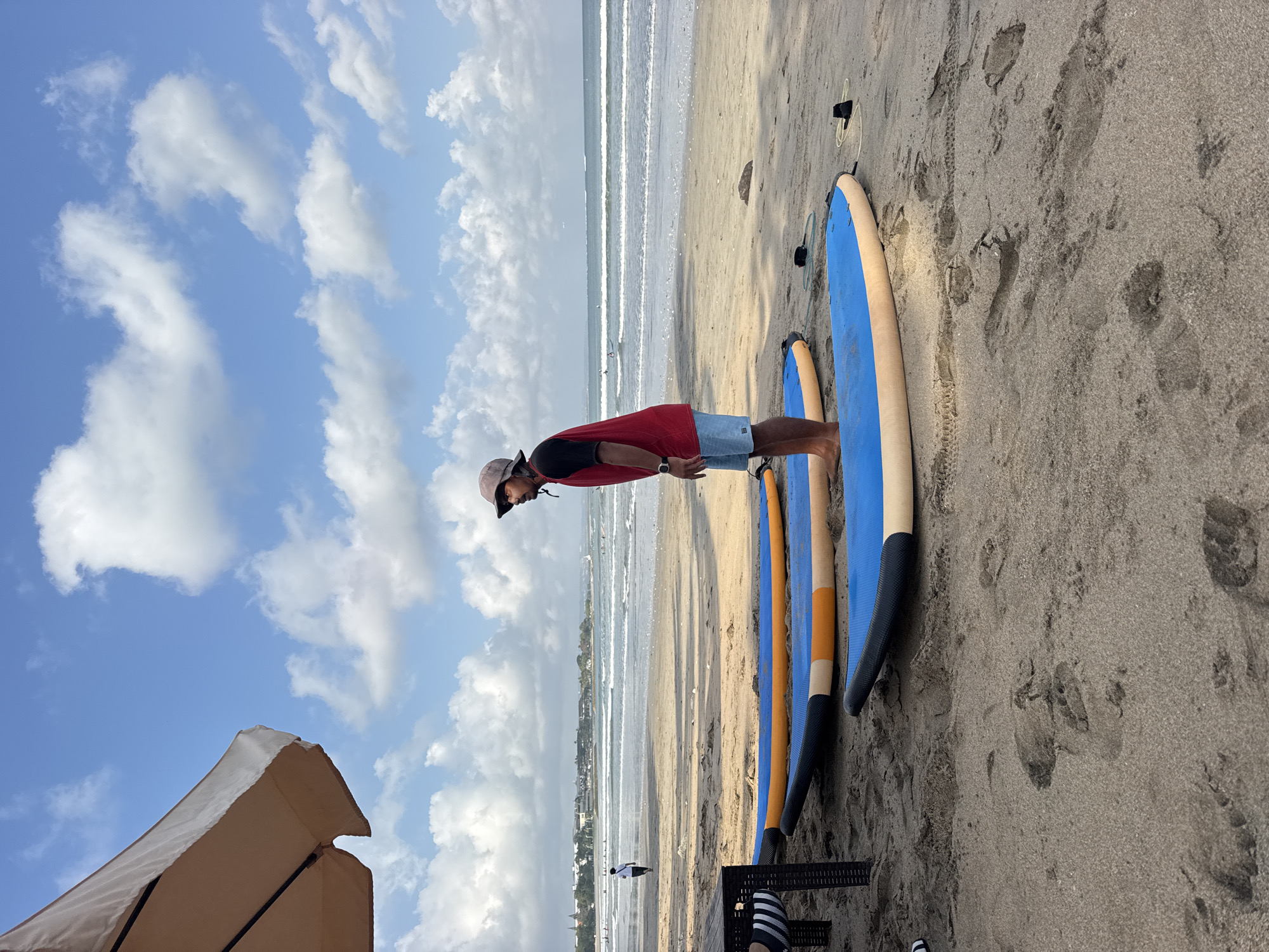 The image depicts a beach scene in Kuta, Indonesia, under a partially cloudy sky. In the foreground, three blue surfboards with orange and black accents lie on the sand. A man wearing a red short-sleeved shirt, blue shorts, a hat, and sunglasses stands between two of the surfboards, facing towards the ocean. His right arm is bent, and his left arm is relaxed at his side. In the mid-ground, the ocean waves roll towards the shore. A few people are visible in the water and on the beach further down. To the left, a portion of a large, light-colored umbrella is visible, casting a shadow on the sand. The sand is textured with footprints and tire tracks, indicating activity on the beach. In the background, a line of buildings and palm trees can be seen along the coastline, typical of the Kuta area.