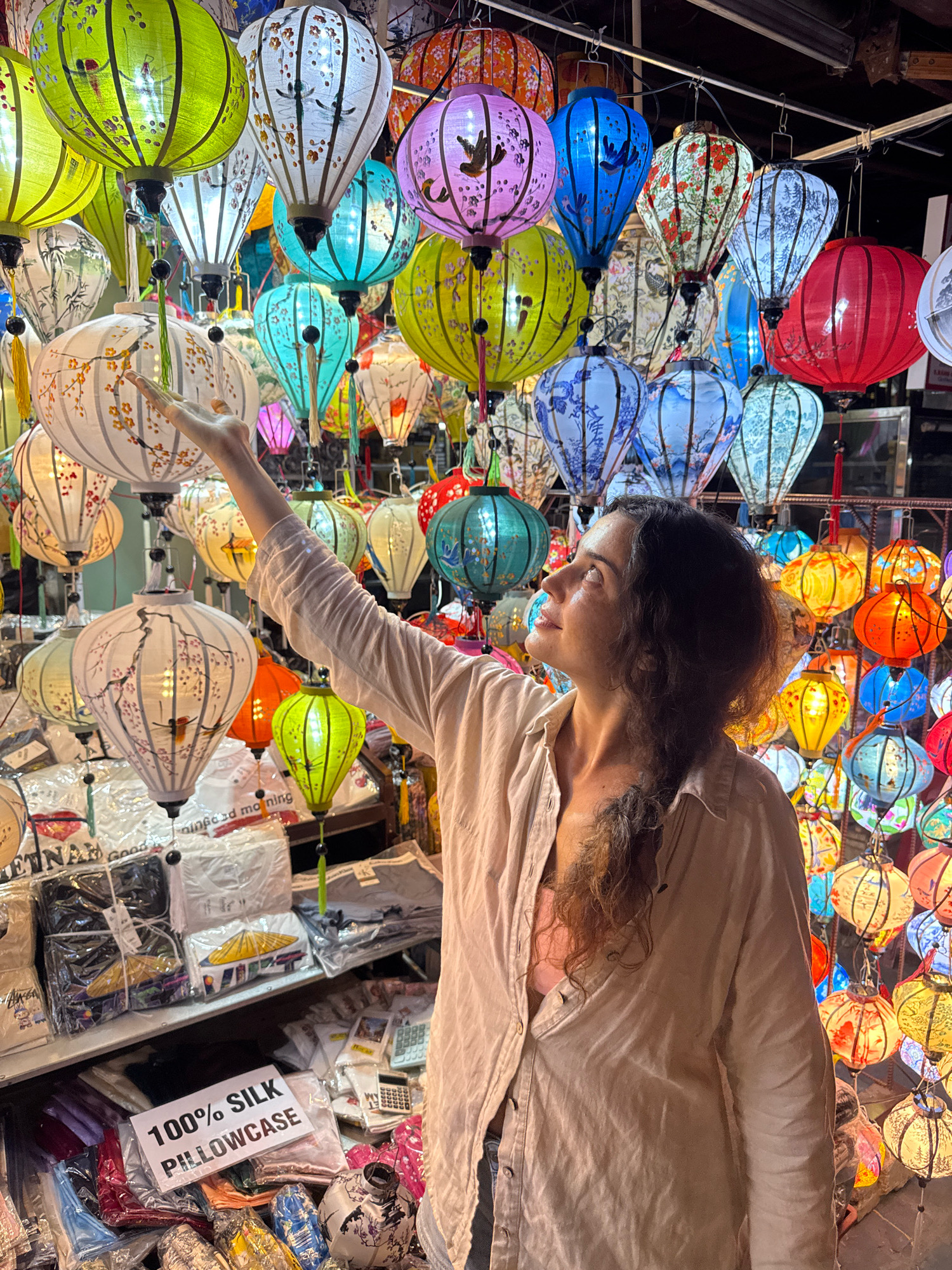 A woman with long, dark, curly hair is standing in a lantern shop in Hoi An, Vietnam. She is wearing a light-colored, unbuttoned shirt over a pink top. Her right arm is extended upwards, with her hand reaching towards a cluster of colorful, illuminated lanterns. The lanterns are of various shapes, sizes, and colors, including green, white, blue, purple, red, and orange, and many are decorated with intricate patterns and designs. The shop appears to be filled with many more lanterns, as well as shelves displaying various goods, including what appear to be textiles and small souvenirs. The lighting in the scene is primarily from the lanterns themselves, casting a warm glow on the woman and the surrounding merchandise. The overall impression is that of a vibrant and bustling market.