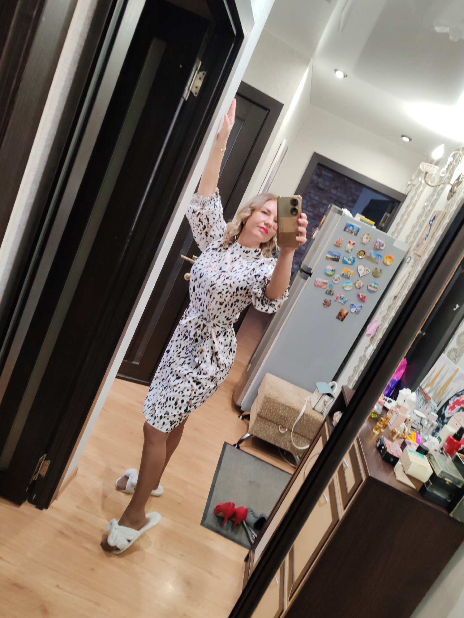 A woman is taking a selfie in a mirror. She has blonde hair and is wearing a patterned dress and white slippers. She is holding a smartphone in her right hand, arm raised up. The background shows a hallway or room with a wooden floor, a closed dark wooden door, a refrigerator covered in magnets, and a dresser with assorted items on top. While this scene is not directly identifiable as Petropavlovsk-Kamchatsky, Russia, there are no elements that would contradict it being located in that city.