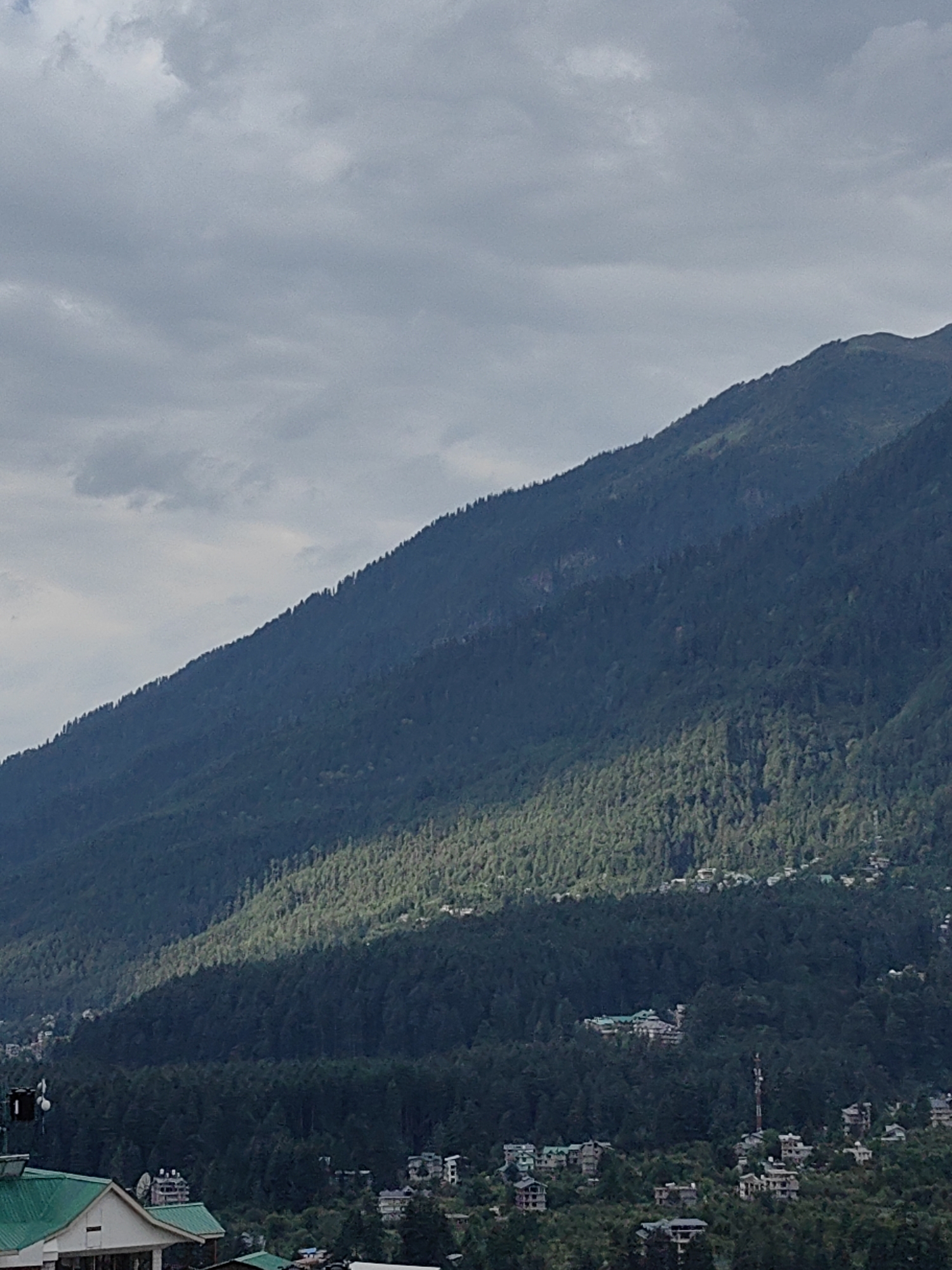 The image depicts a mountainous landscape with a town nestled at its base. The sky is overcast with a mix of dark and lighter clouds. A large, densely forested mountain slope dominates the scene, with a diagonal band of sunlight illuminating a portion of the trees. Several small buildings and structures are scattered across the lower slopes and the valley floor, suggesting a residential or tourist area. The architecture of the buildings is varied, with some appearing to be multi-story and others more traditional. Vegetation is abundant, with the mountain covered in a dark green forest and smaller trees and foliage visible in the town. The overall impression is one of a scenic, natural environment with human habitation. The specific mention of Vashisht, India, places the scene in a known tourist destination in the Himalayas, often characterized by such mountain views and built-up areas.
