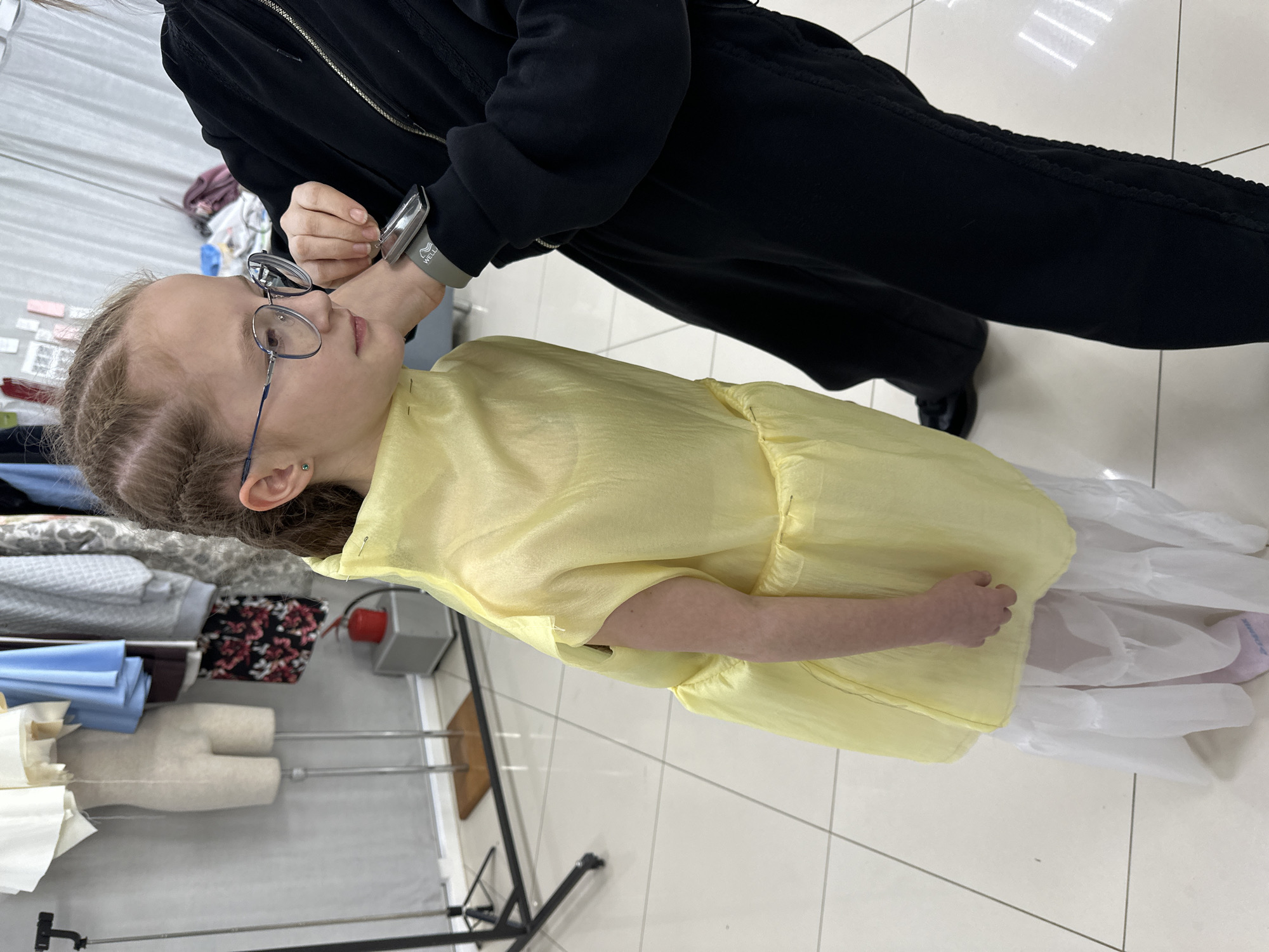 A young girl with braided hair and glasses is being fitted for a yellow dress by an individual wearing a black tracksuit. The location appears to be a tailor's workshop, with fabric, a dress form, and tools visible in the background. The setting is illuminated by overhead fluorescent lighting. There is no confirmation that this image was taken in Votkinsk, Russia.