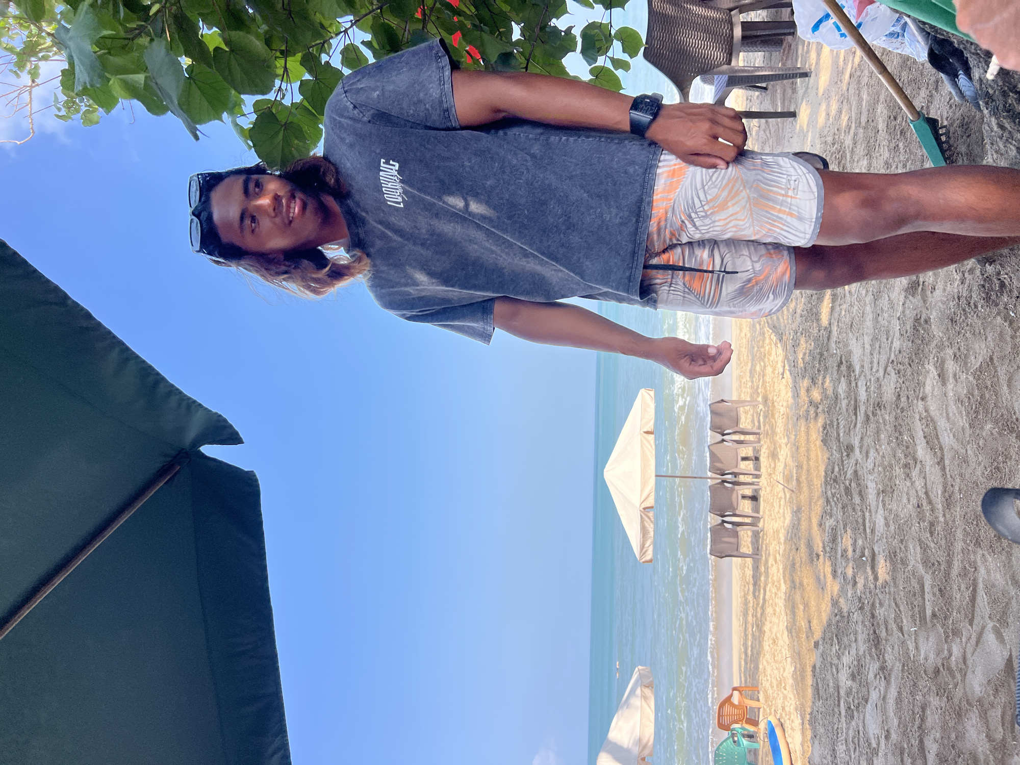 A young man with long, wavy, dark hair and sunglasses perched on his head stands on a sandy beach. He is wearing a charcoal-colored, distressed-look t-shirt with white text and patterned swim shorts featuring palm leaves in orange and gray. He has a watch on his left wrist. Behind him, the turquoise ocean meets a clear blue sky. Several beach umbrellas and rows of empty chairs are set up on the sand. In the foreground, sand is visible, with a green rake and a white plastic bag to the right. The overall scene suggests a relaxed atmosphere at a beach in Kuta, Indonesia.