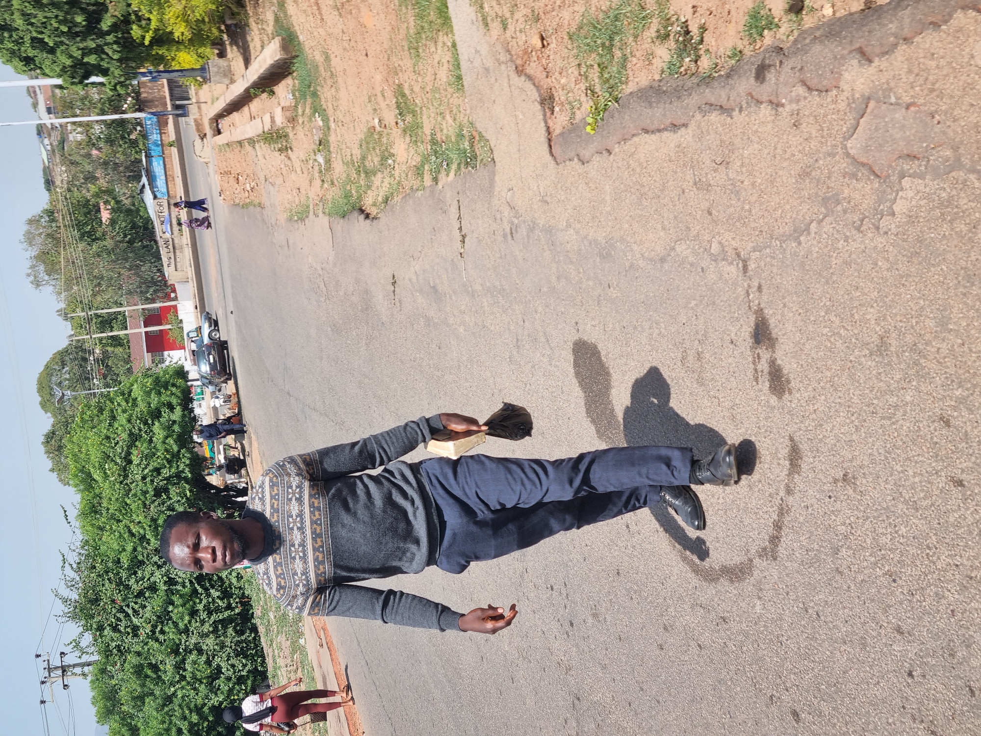 A man stands on a paved road in Jos, Nigeria. He wears a gray patterned sweater, navy trousers, and black shoes, holding a small object in his left hand. Behind him, there is a busy street with cars and pedestrians. Lush green trees and vegetation line the roadside. The environment appears to be a sunny day in an urban setting.