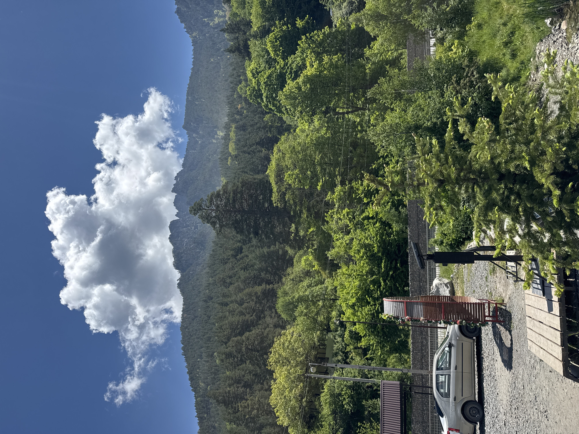 The image captures a scenic view of a mountainous region with dense evergreen forests blanketing the slopes under a bright blue sky with scattered white clouds. In the foreground, a gravel lot is visible with a light-colored sedan parked on the left. Adjacent to the car is a red, cylindrical structure decorated with flowers, possibly an art installation or a decorative element. A wooden platform is also present in the foreground, and a lamppost stands nearby. A low stone wall runs horizontally across the middle ground, separating the gravel area from a grassy slope covered in lush green trees. The background is dominated by the imposing, forested mountain range, its peaks sharp against the sky. The image appears to be taken in the city of Teberda, Russia, a location known for its natural beauty and mountainous terrain.