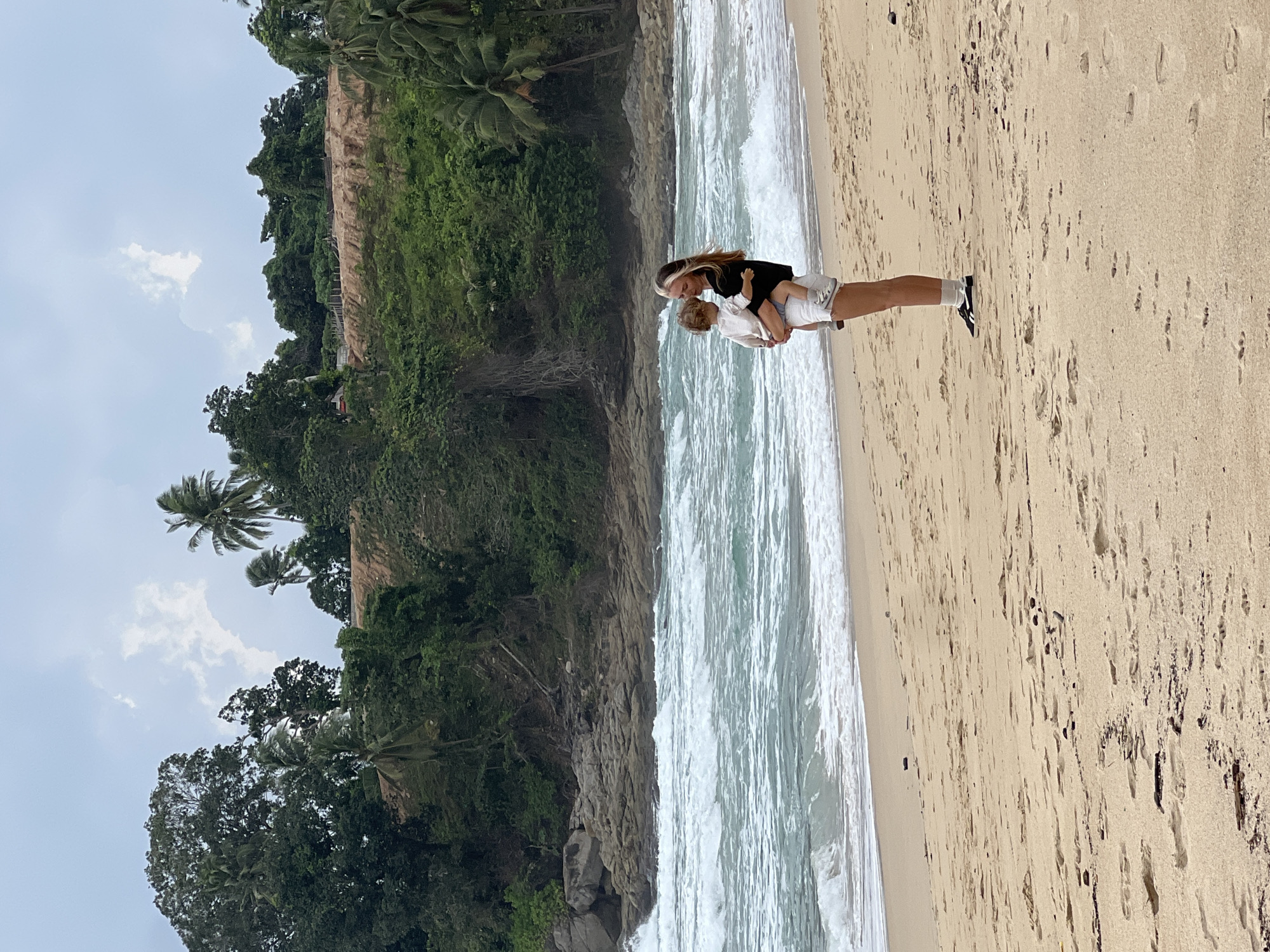 A young woman with blonde hair is standing on a sandy beach in Choeng Thale, Thailand, holding a toddler. The woman is wearing a black top and white shorts, and the toddler is wearing a white shirt and light-colored shorts. They are facing towards the ocean, which has moderate waves. In the background, there is a lush green hillside with palm trees and rocky outcrops leading down to the water. The sky is overcast.