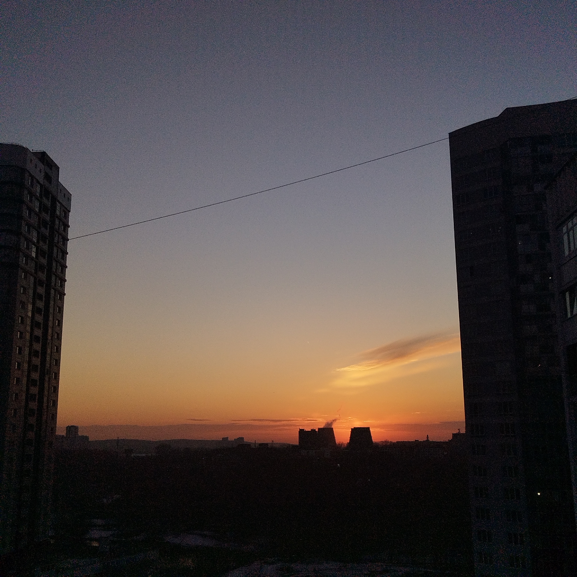 An elevated long shot depicts an urban landscape in Yekaterinburg, Russia, at either sunrise or sunset. The sky transitions from a deep indigo at the top to vibrant orange and yellow hues near the horizon. A thin, horizontal cloud streak is present in the lower right sky. Two tall, multi-story buildings, appearing as dark silhouettes, frame the central view, one on the far left and one on the far right. A single, thin dark wire stretches horizontally across the sky, connecting approximately the upper-middle portions of these two foreground buildings. In the distant mid-ground, a dark, silhouetted urban horizon reveals smaller buildings and two prominent, large, industrial-type structures, possibly cooling towers or chimneys, one emitting a plume of illuminated smoke or steam. The ground level is dark and indistinct, showing mottled patches of what appears to be snow or ice. No individuals or discernible actions are present.