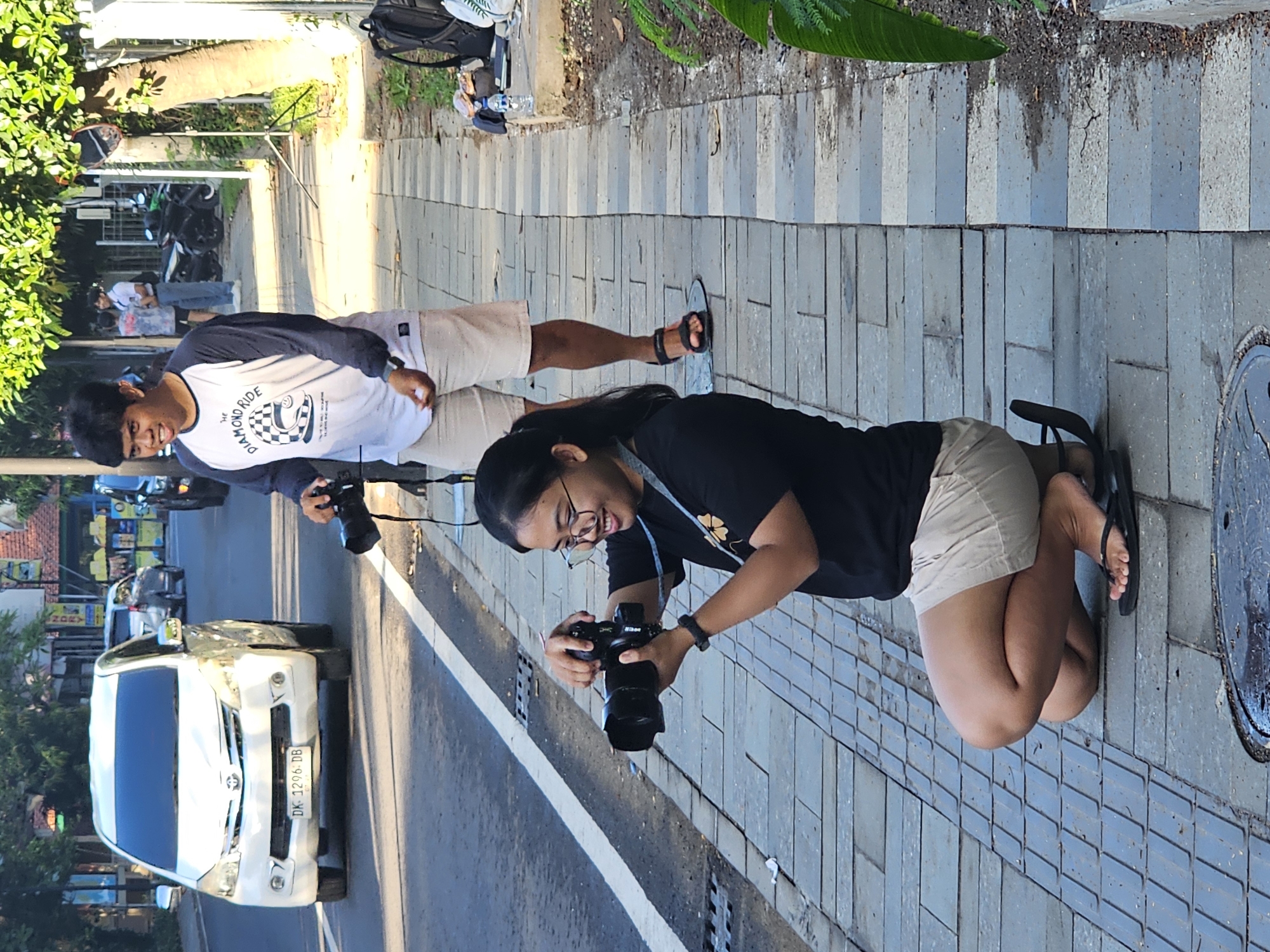 Two people stand and kneel on a brick sidewalk next to a street in Denpasar, Indonesia. The person standing, presumably a male, is in a white and navy long-sleeve shirt with a graphic on the front, light-colored shorts, and black sandals. They are holding a black camera in their hands. The other person, presumably a female, is kneeling and holding a similar camera. They are wearing a black t-shirt with a small graphic on the front, light-colored shorts, glasses, and black sandals. A light-colored van passes on the street, with a license plate clearly visible that reads "DK 1296 DB". Trees, buildings, and other vehicles are visible across the street. A round metal manhole cover is partially visible near the kneeling person.