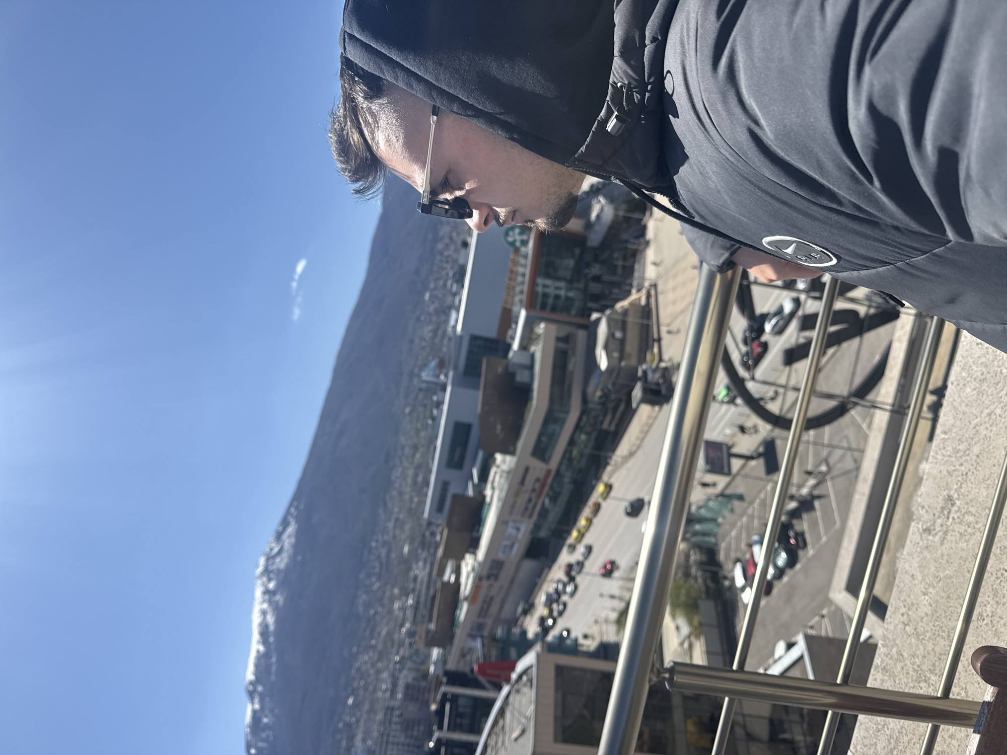 A man in a black hooded jacket stands on a balcony, looking down at the city of Sofia, Bulgaria. He is wearing sunglasses. The balcony has a metal railing. Below, there is a street with cars and buildings. In the background is a mountain range. The sky is blue and clear.