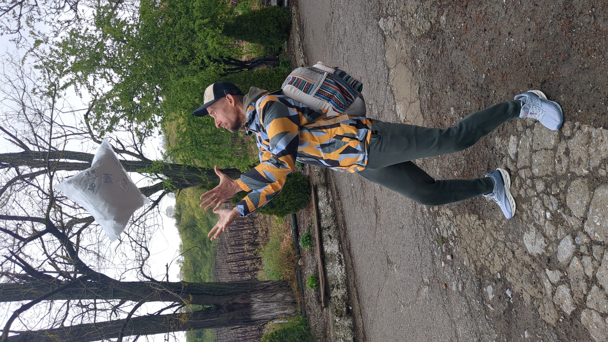 A man in a yellow and black patterned jacket, dark green pants, and light blue sneakers is depicted in an outdoor scene. He's wearing a baseball cap and has a backpack on. The man is positioned with his arms outstretched as if attempting to catch a white plastic bag floating in the air. The background includes bare tree branches and green foliage, suggesting a park or wooded area. The ground appears to be a cracked asphalt path. Based on the filename, the location is Kiparisnoe, Ukraine.