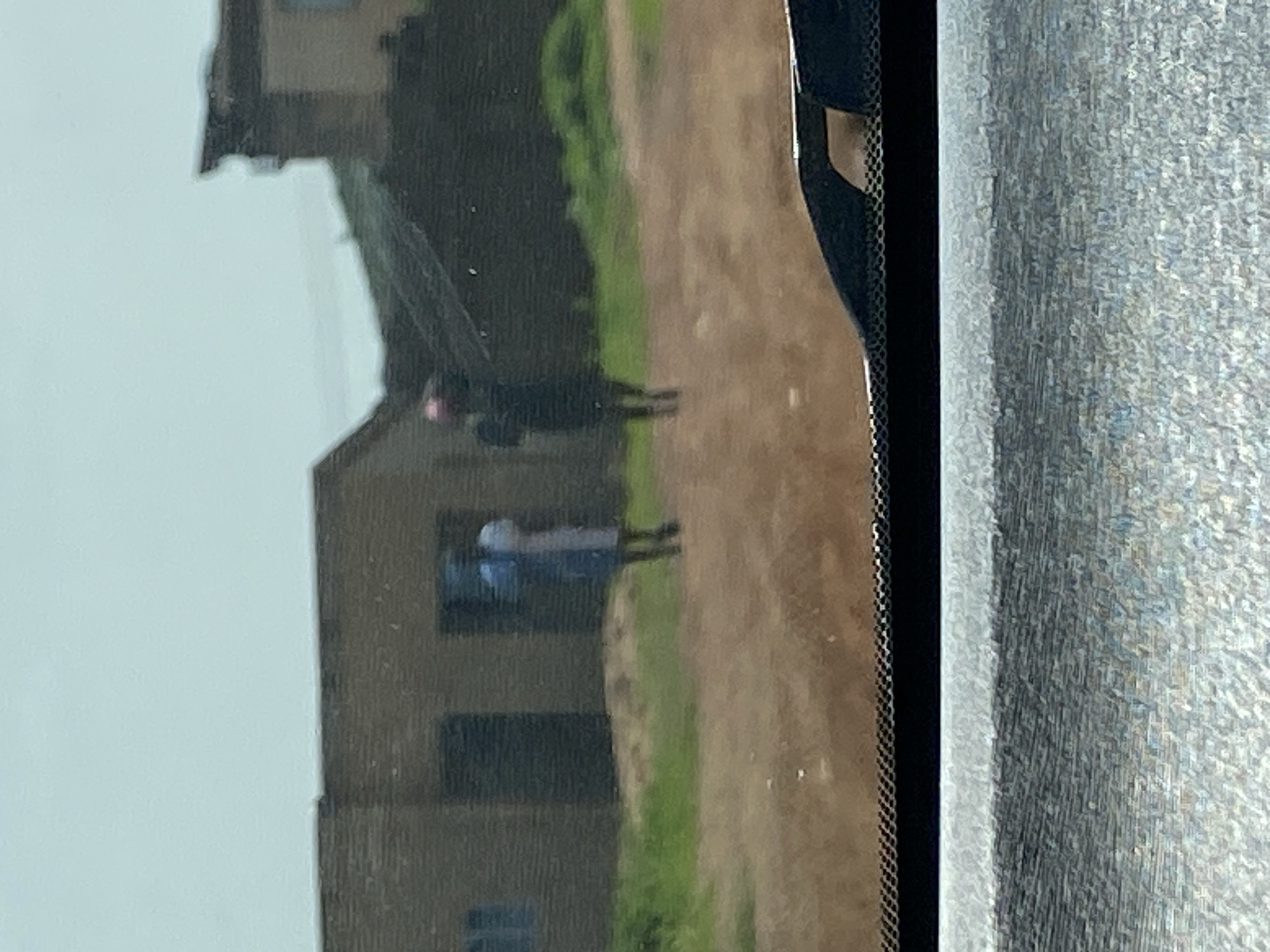 Two individuals are visible in the mid-ground of the image, standing on a dirt path in front of a low-rise, brick building. The person on the left appears to be wearing a blue garment. The person on the right is wearing a dark garment and what may be a pink head covering. The building behind them has at least two dark rectangular openings, possibly windows or doorways. To the right of this building, another structure with a darker, slanted roof is partially visible. The environment is rural, with patches of green grass interspersed with the dirt path and building. The foreground shows the edge of a vehicle's interior, suggesting the image was taken from inside a car. The sky is overcast.