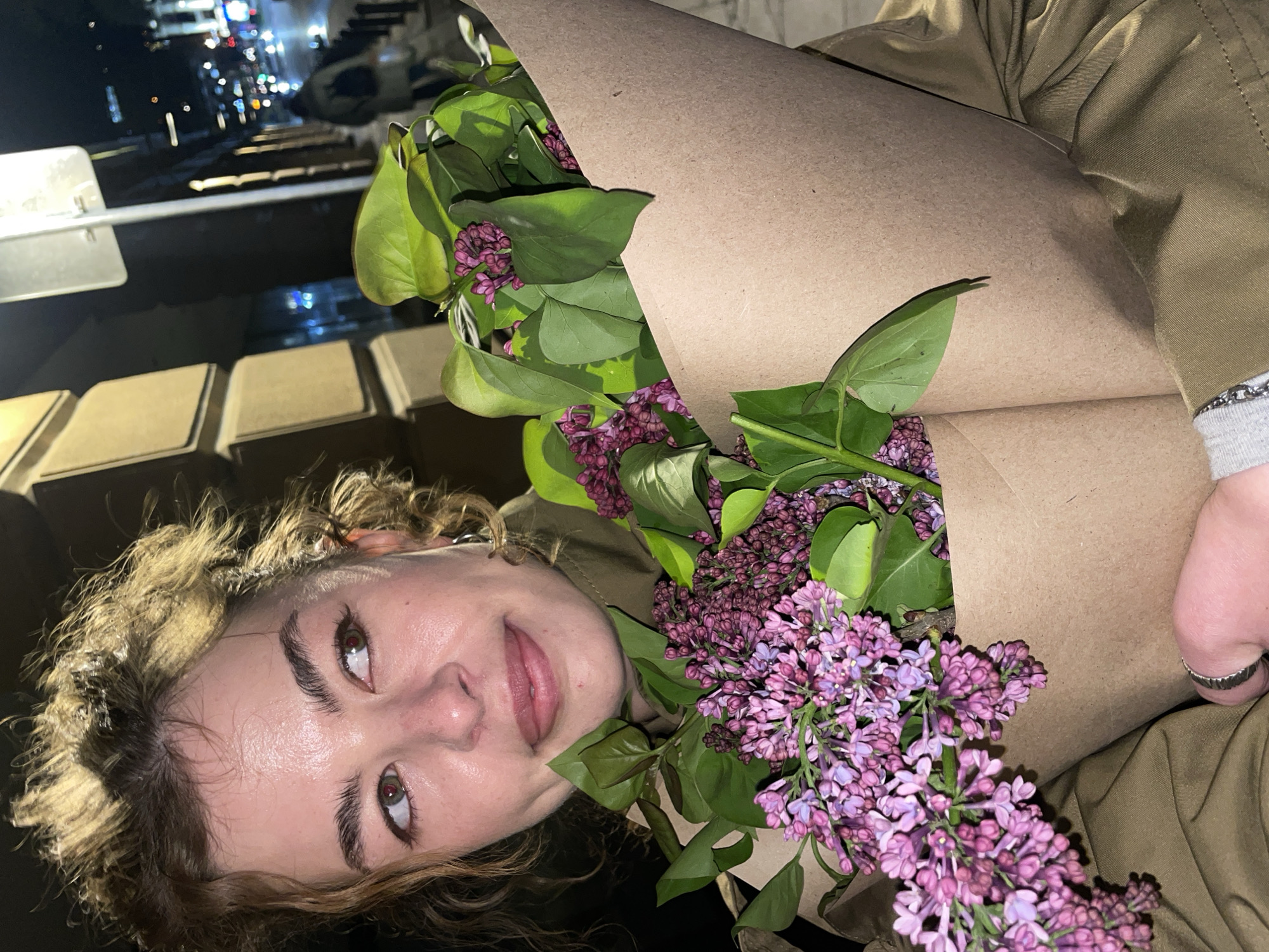 A woman is holding two bouquets of purple lilacs wrapped in brown paper. The woman is Caucasian with curly light brown hair, light skin, and light green eyes. She wears a khaki-colored jacket. The location is the city of Tbilisi, Georgia, at night, based on the backdrop which includes streetlights and other blurred lights. The bouquets are the main focus, with the woman holding them in front of her.