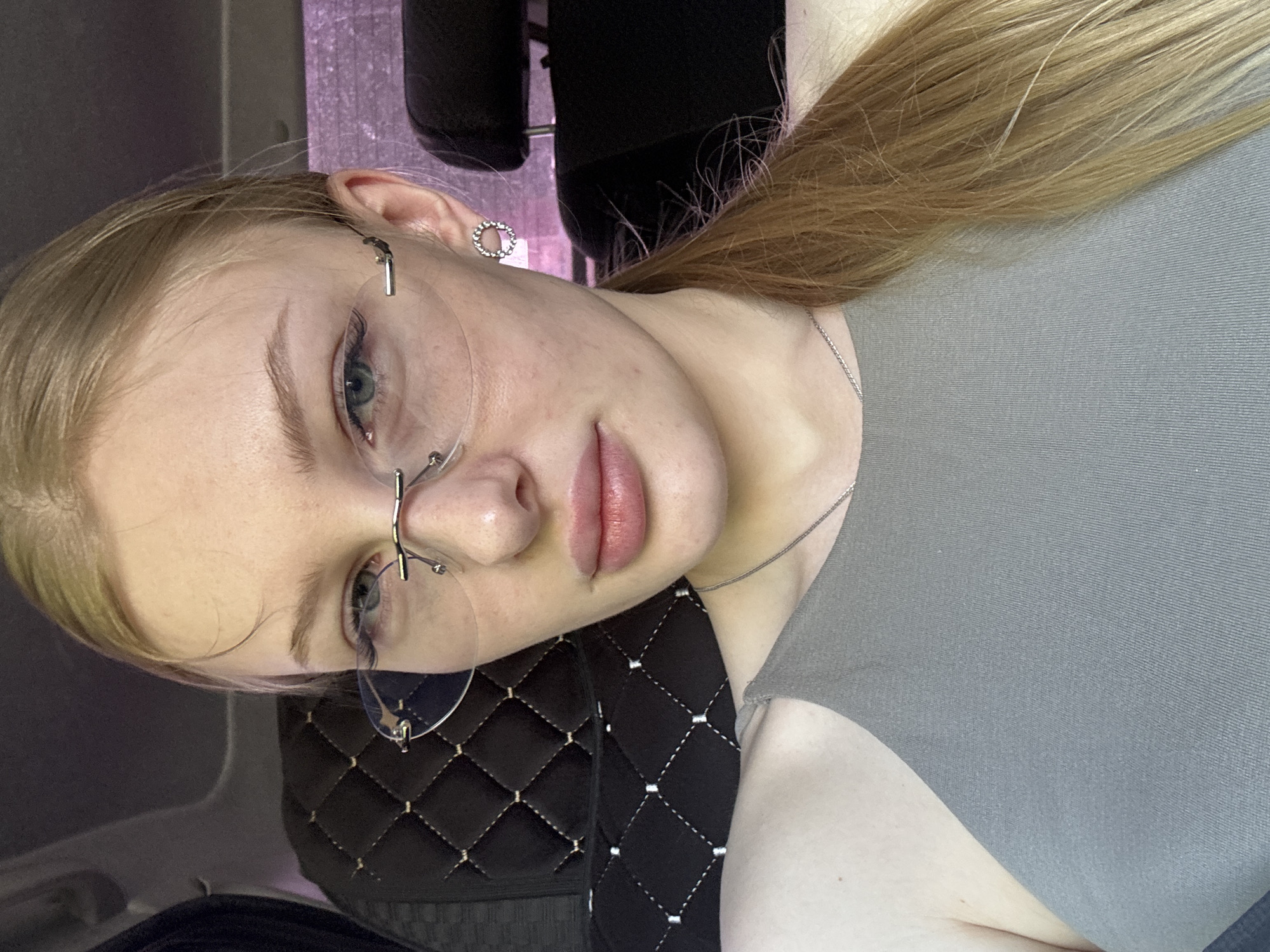 A young, light-skinned woman with long blonde hair pulled back in a ponytail is the primary subject of the image. She is wearing rimless glasses and a gray top. A silver necklace is visible around her neck, and she has a circular earring in her left ear. The background appears to be the interior of a car, with a black quilted seat cover visible. A headrest is also visible behind her. The setting is likely in Perm, Russia, although specific landmarks or street signs are not visible in the photograph to confirm this definitively.