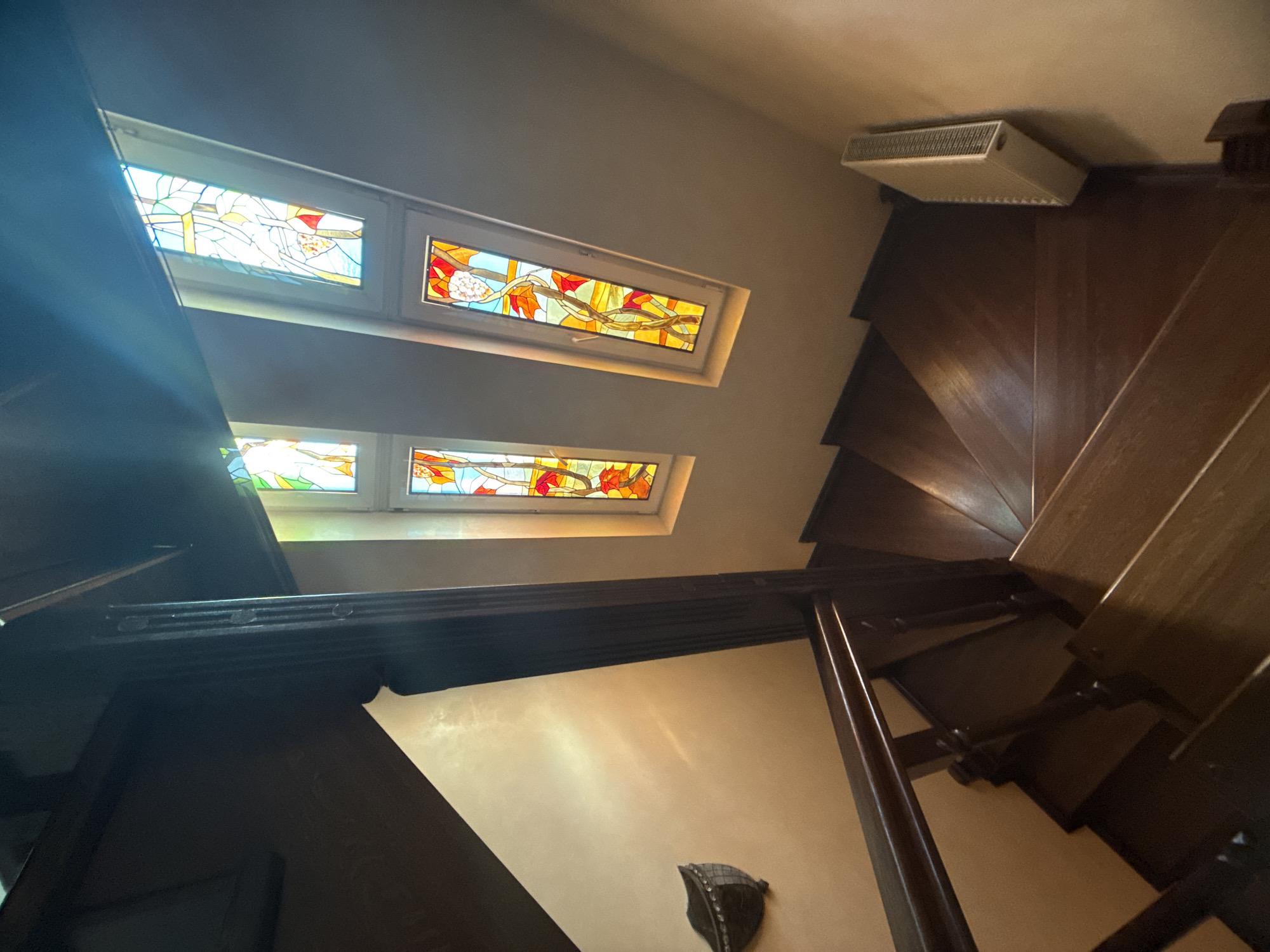 The image shows a view looking down a dark wooden spiral staircase. The staircase has a wooden railing with carved posts. Two narrow, vertical stained-glass windows are visible at the top of the staircase, letting in colorful light. One window shows an abstract design with yellow, red, and blue glass, and the other shows a vine with orange and yellow leaves and red flowers. To the left of the staircase, a dark sconce is mounted on a cream-colored wall. A white radiator is mounted on the wall to the right of the bottom of the staircase. The environment appears to be an indoor staircase in a residential or public building in the city of Yukki, Russia. There are no people visible in the image.