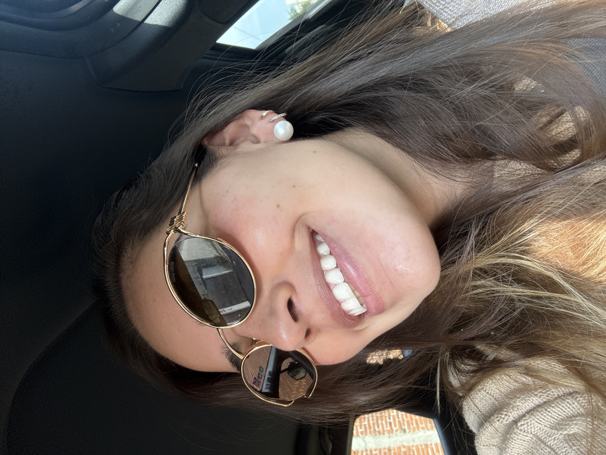 A selfie-style image captures a young woman inside a car, smiling brightly. Her head is tilted significantly to the right, with her long, dark brown hair, featuring lighter streaks, flowing around her shoulders. She wears gold-framed, round sunglasses, and a large pearl stud earring along with a smaller, diamond-encrusted ear cuff on her visible ear. The brand "MIU MIU" is visible on the sunglasses frame. She is wearing a light-colored, possibly beige or cream, knitted top.
Bright sunlight streams into the car from an overhead window or sunroof, illuminating her face and hair. The car's dark interior headliner is visible in the background. Reflections in her sunglasses show outdoor elements, including what appears to be a brick wall and bright sky, consistent with the brick wall visible through the car window in the lower right background. Her broad smile reveals her upper teeth, conveying a cheerful emotion. The scene suggests a bright, sunny day, likely in San Bernardino Tlaxcalancingo, Mexico.