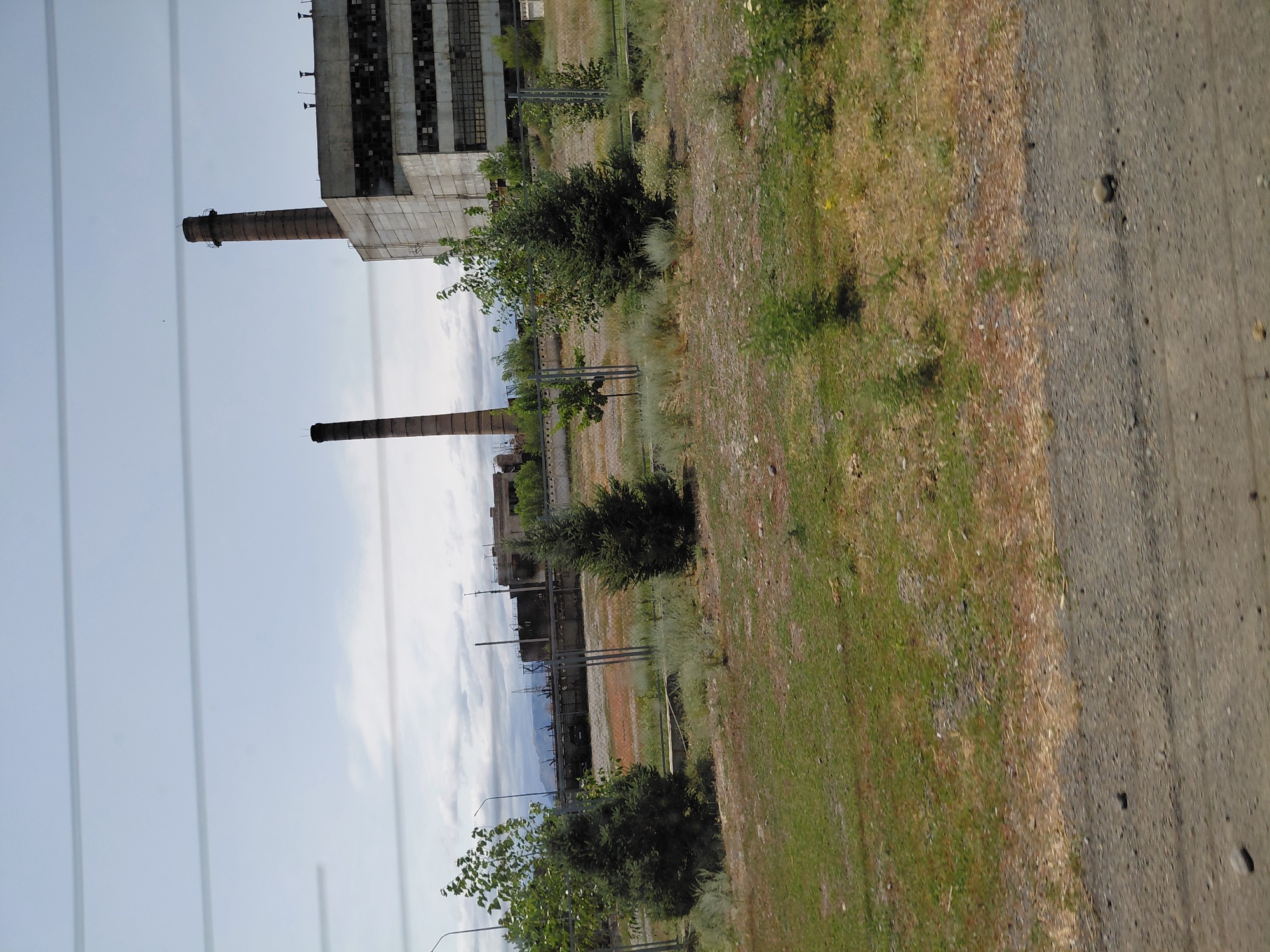 The image captures a view of an industrial area under a cloudy sky. Dominating the scene are two large, brick chimneys, one taller than the other, situated next to a large, rectangular, concrete industrial building with broken windows. The building appears weathered and abandoned. In the foreground, there is a patch of dry, brown grass interspersed with green patches and scattered stones, indicating a dry, possibly arid environment. Several small, conical evergreen trees and other sparse vegetation are scattered throughout the area, some behind a metal fence. The overall impression is one of an abandoned industrial site, likely in a somewhat remote or neglected part of a city. The geographical context provided by the prompt suggests this is an industrial facility in Бұлақты, Kazakhstan.