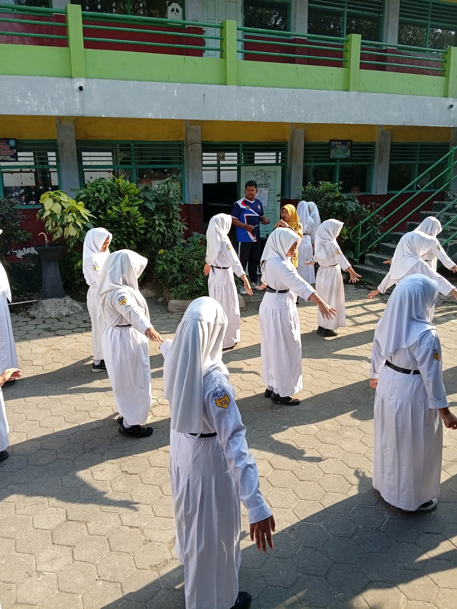 In Lawang, Indonesia, a group of female students in white uniforms and hijabs are engaged in a physical activity outdoors, likely a form of exercise or dance. They are positioned on a paved courtyard with a hexagonal pattern. A male instructor, wearing a blue and white t-shirt and dark pants, stands near a building and observes them. The building, with its distinct green, red, and yellow facade and balconies, appears to be a school. Lush green foliage is visible in front of the building, and a set of stairs leads up to an entrance on the right side of the image. The sunlight casts long shadows on the ground, indicating it is daytime.