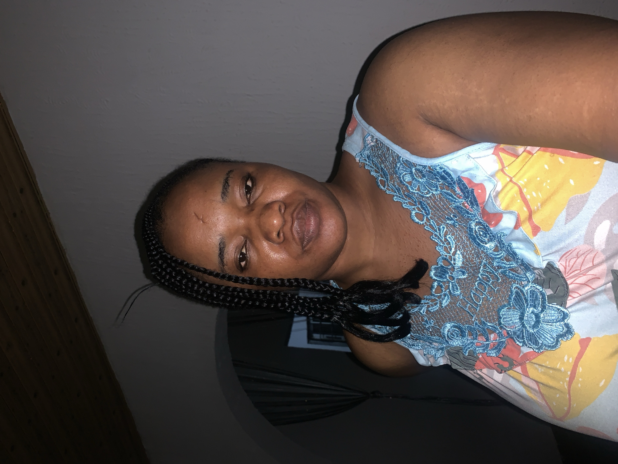 A medium shot captures a Black woman with braided hair and a floral print dress. The woman is positioned in a room with a dark, textured ceiling, and a plain grey wall. Her body is angled slightly away from the camera, and she is looking directly at the viewer with a neutral expression. The woman has a small scar on her forehead and is wearing a blue lace-trimmed dress with a vibrant floral pattern. The background appears to be indoors, with a hint of an arched doorway to the left, partially obscured by darkness and curtains. The overall lighting is dim, casting shadows across the scene, suggesting an indoor setting in Barakin Akawo, Nigeria.