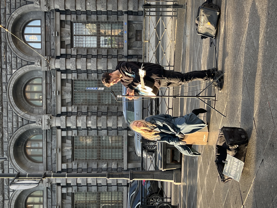 This image captures a street performance scene on a sunny day in Saint Petersburg, Russia, on a sidewalk adjacent to the Russian Museum complex, likely on Inzhenernaya Street or in the vicinity of Arts Square.
In the center, two individuals are present. A male musician stands on the right, dressed in a black hoodie, black cargo pants, and black sneakers. He holds an acoustic guitar, looking down at it, potentially tuning or adjusting it. A black microphone on a tripod stand is positioned in front of him. To his right, a small, black "CUBE" amplifier rests on the grey pavement. To the left, a female musician is seated on a light-colored wooden cajon. She wears a long, dark blue or grey coat and dark shoes, with long blonde hair illuminated by strong sunlight from the left. She is looking towards the male musician with a mild smile.
In the foreground, a black fabric container functions as a donation box, holding a white sign with a social media handle (@milicajamin), a Russian phone number (8-900-623-60-40), and text indicating "Donation (Чаевые, payment methods)."
The background features a large, imposing grey stone building characterized by heavy rustication on its lower levels, multiple arched windows on its upper floor, and rectangular, barred windows below. This architecture is consistent with the Russian Museum of Ethnography or the Mikhailovsky Palace of the State Russian Museum. A dark streetlight pole stands on the far left. A low black metal fence separates the sidewalk from the street in the background, where several vehicles, including a dark SUV and a black sedan, are visible. The ground is grey pavement, reflecting sunlight and displaying long shadows, indicative of either morning or late afternoon lighting conditions.
