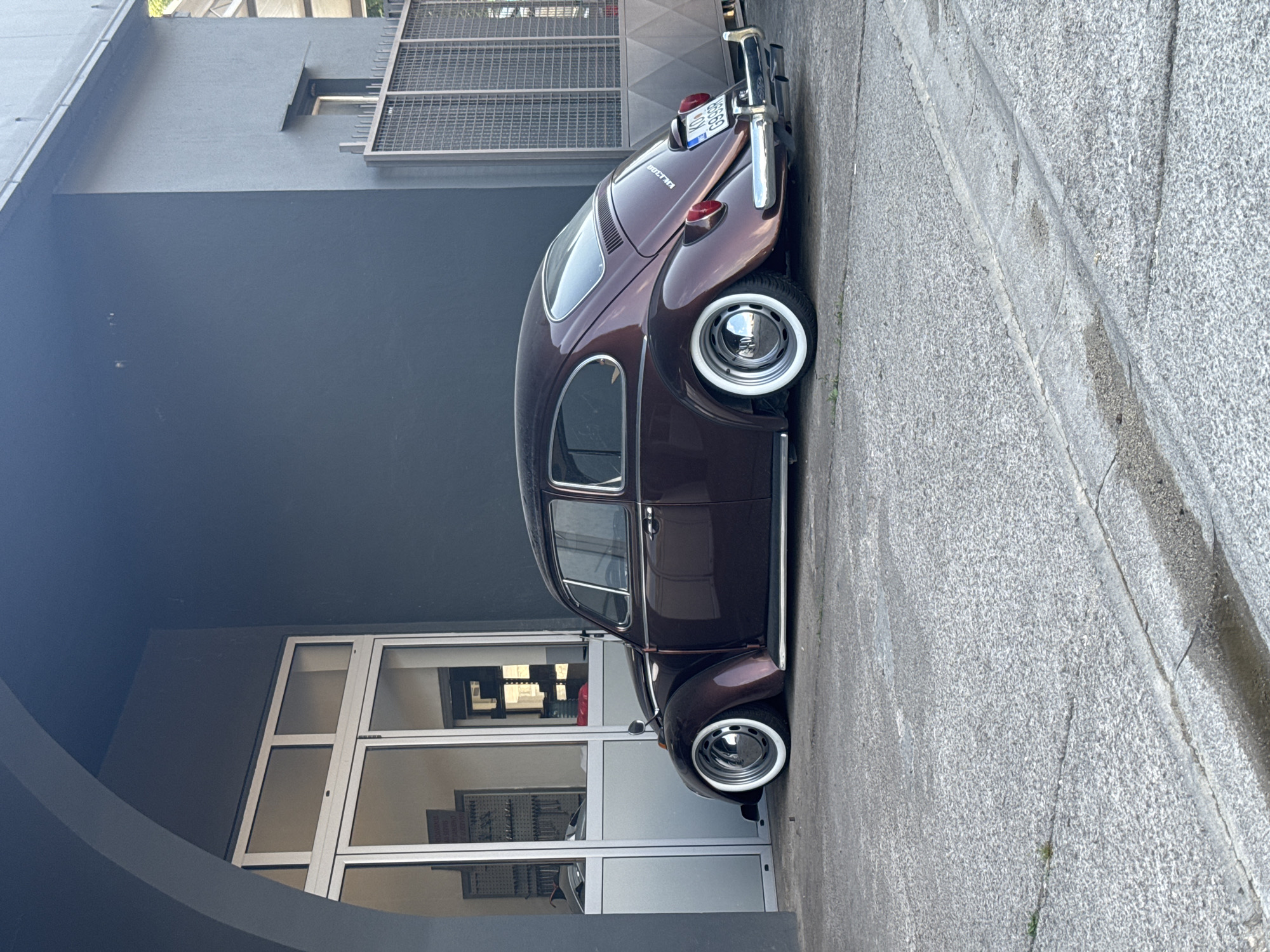 A dark metallic brown, lowered Volkswagen Beetle is parked on a concrete surface in front of a dark gray building. The building has a large glass door and window section on the left, revealing an interior with shelves and tools. To the right of the building is a metal grate fence. The car has whitewall tires and chrome hubcaps. The license plate on the rear of the car reads "K DV G996B". The scene appears to be in an industrial or commercial area. The time of day appears to be daylight, with shadows indicating the sun is to the right. The overall impression is one of a well-maintained, classic car in a somewhat utilitarian setting.