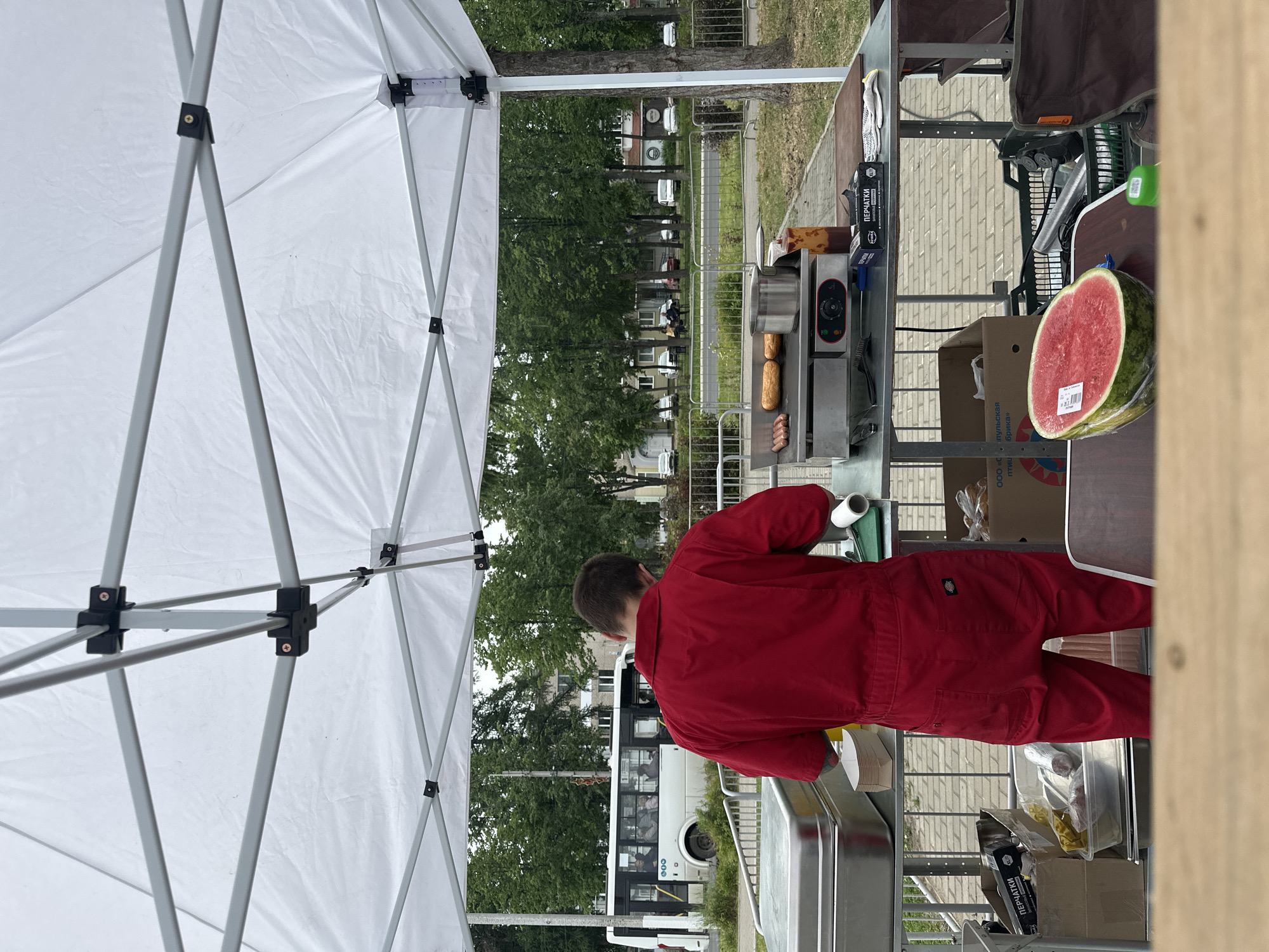 A person in a red jumpsuit is standing behind a food cart under a white tent. They are facing a grill with hot dogs and buns. In front of the grill are various containers and boxes. To the left of the food cart, a white bus is parked on a street. The background shows trees, buildings, and more vehicles. On a table in the foreground, there is a half of a watermelon. The scene appears to be an outdoor event or market in the city of Chaikovsky, Russia.