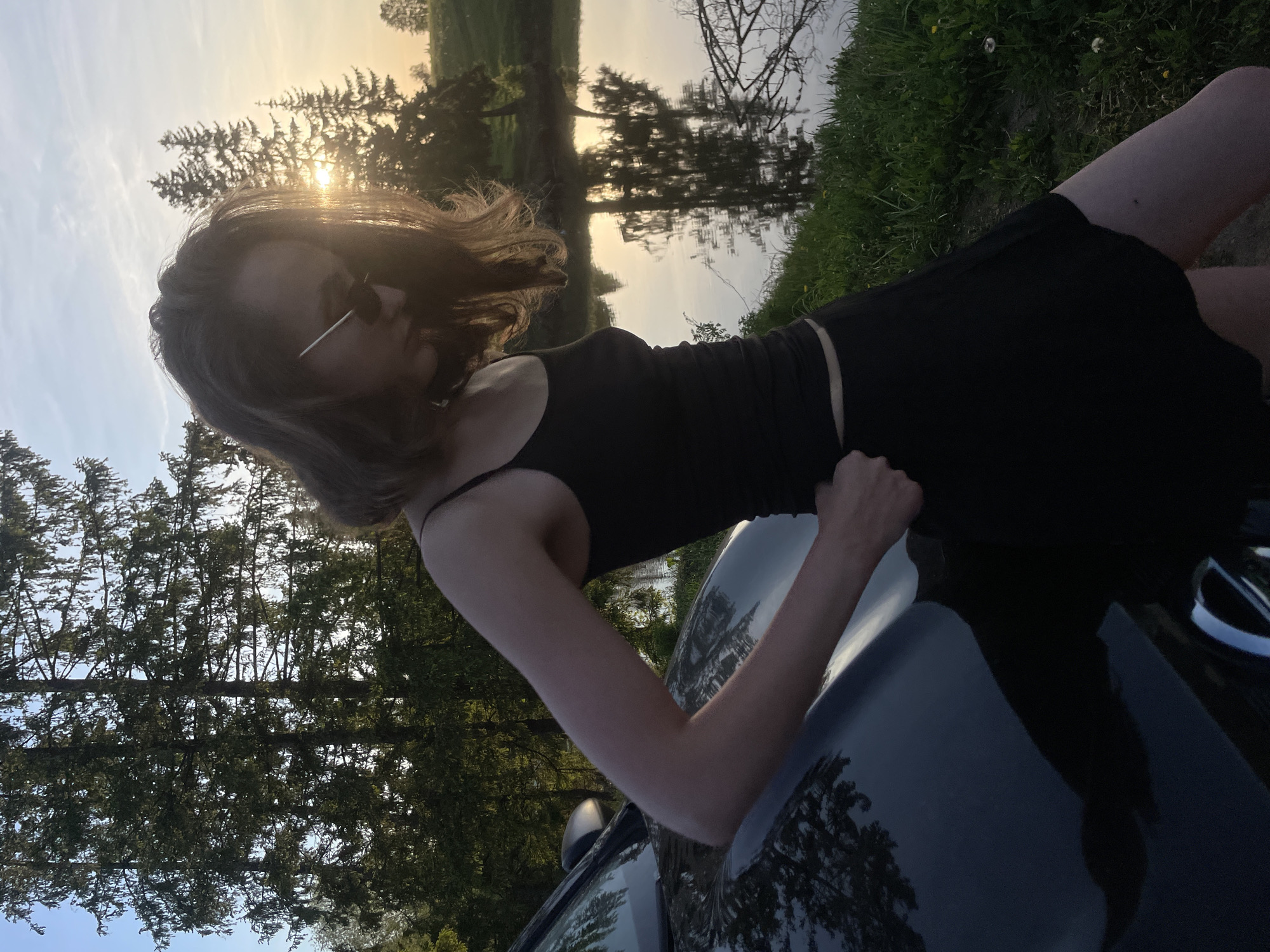 A young woman is leaning on a dark-colored car, likely a sedan, at what appears to be an outdoor location. She is wearing a black tank top and a black skirt. Her hair is shoulder-length and windswept. The sun is setting in the background, with trees and what seems to be a body of water. The reflection of trees is visible on the car's surface. The location seems to be in Podshivalovo, Russia, as the query mentions the location. The woman's pose and the lighting suggest a candid or posed shot during the late afternoon or early evening.